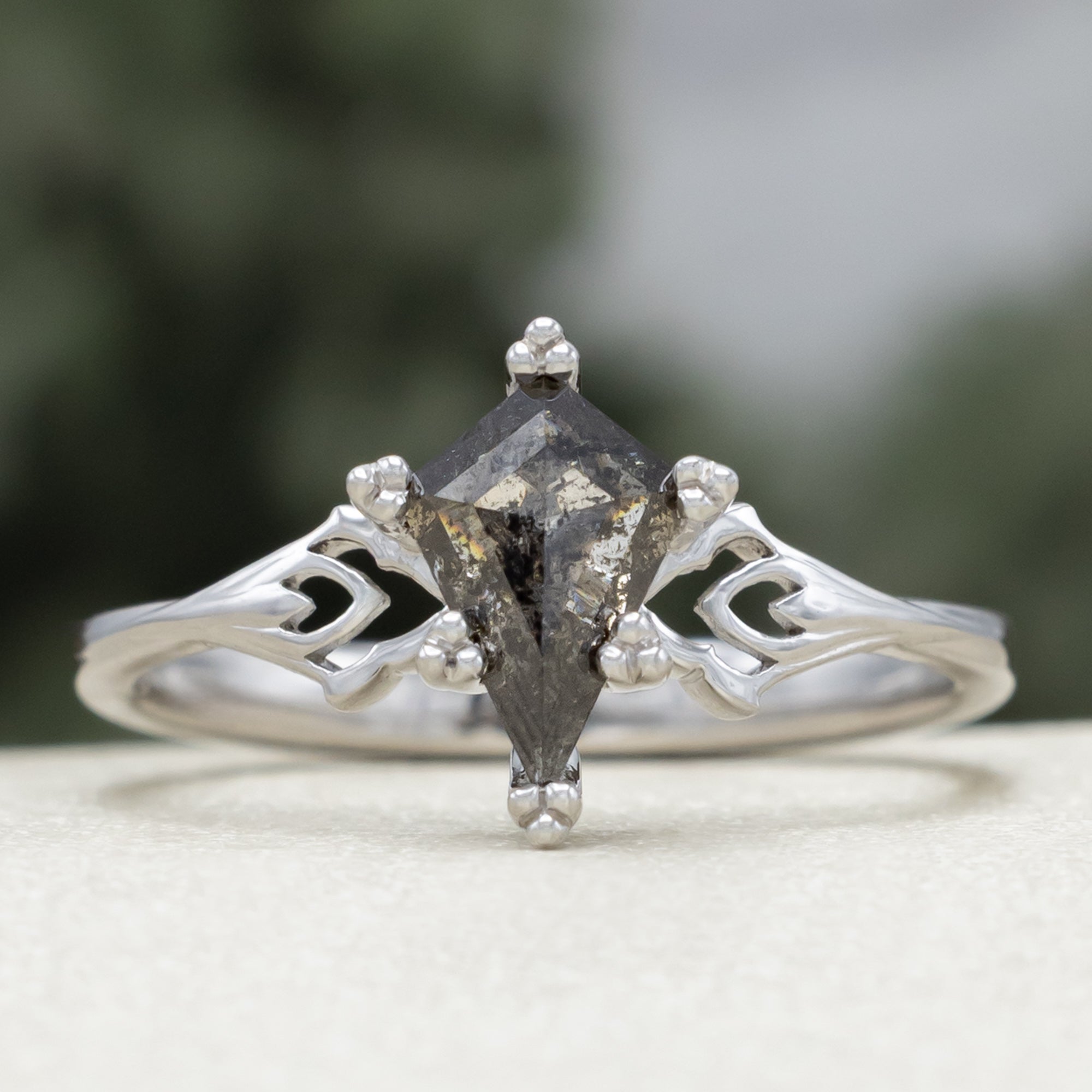 White gold ring with a kite cut salt & pepper diamond and intricate filigree against a blurred background