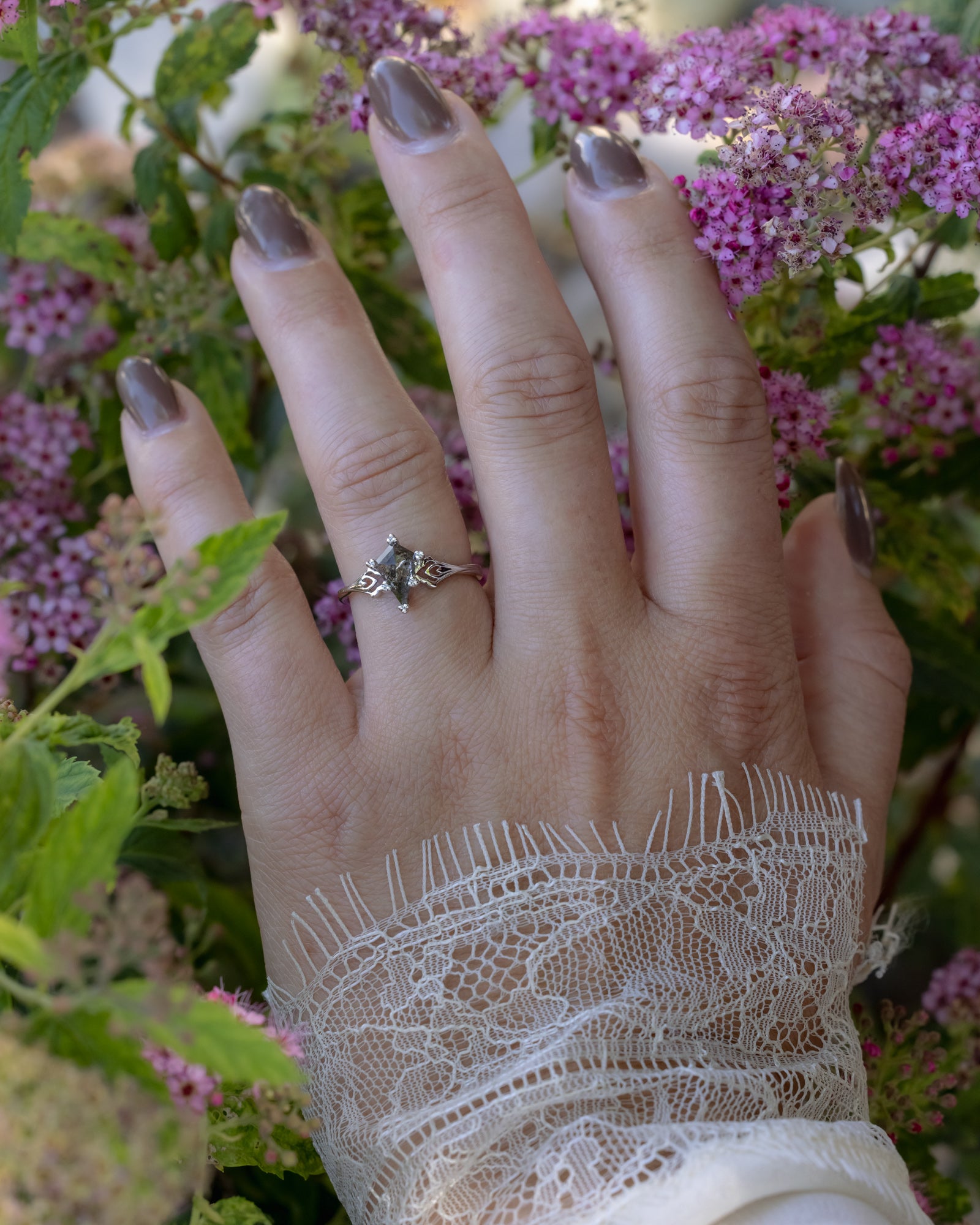 Hand wearing a salt & pepper diamond ring with lace sleeve against a floral background