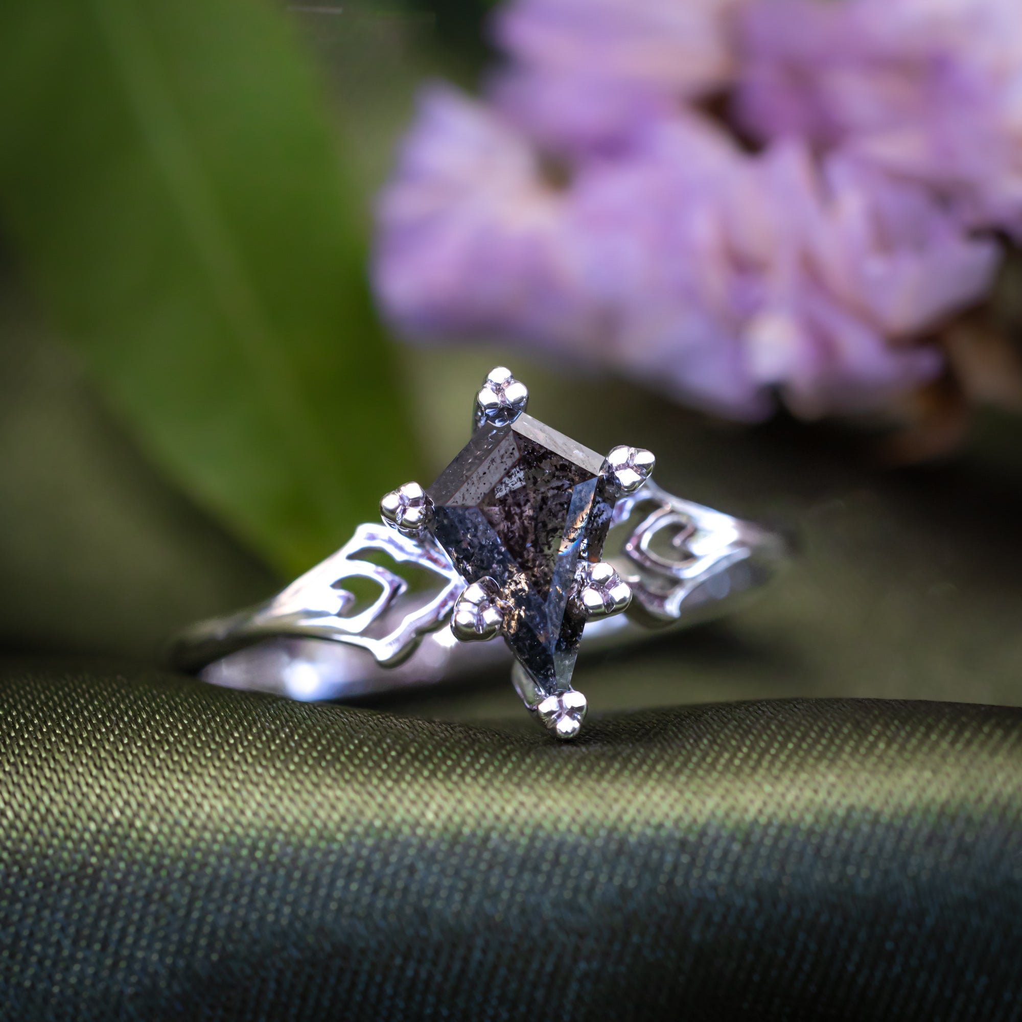 White gold ring with a kite cut salt & pepper diamond on a textured surface with blured flowers in the background
