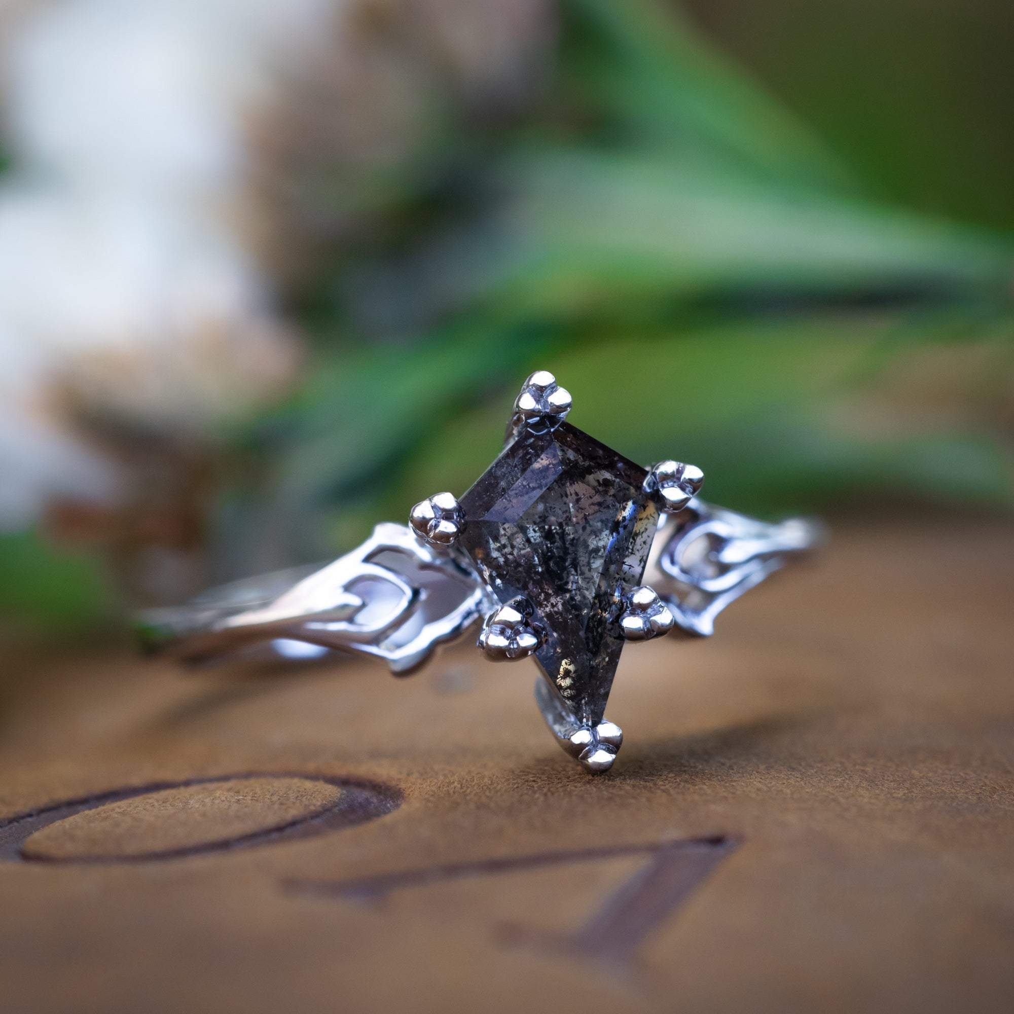 White gold ring with a kite cut salt & pepper diamond on a textured surface with green leaves in the background