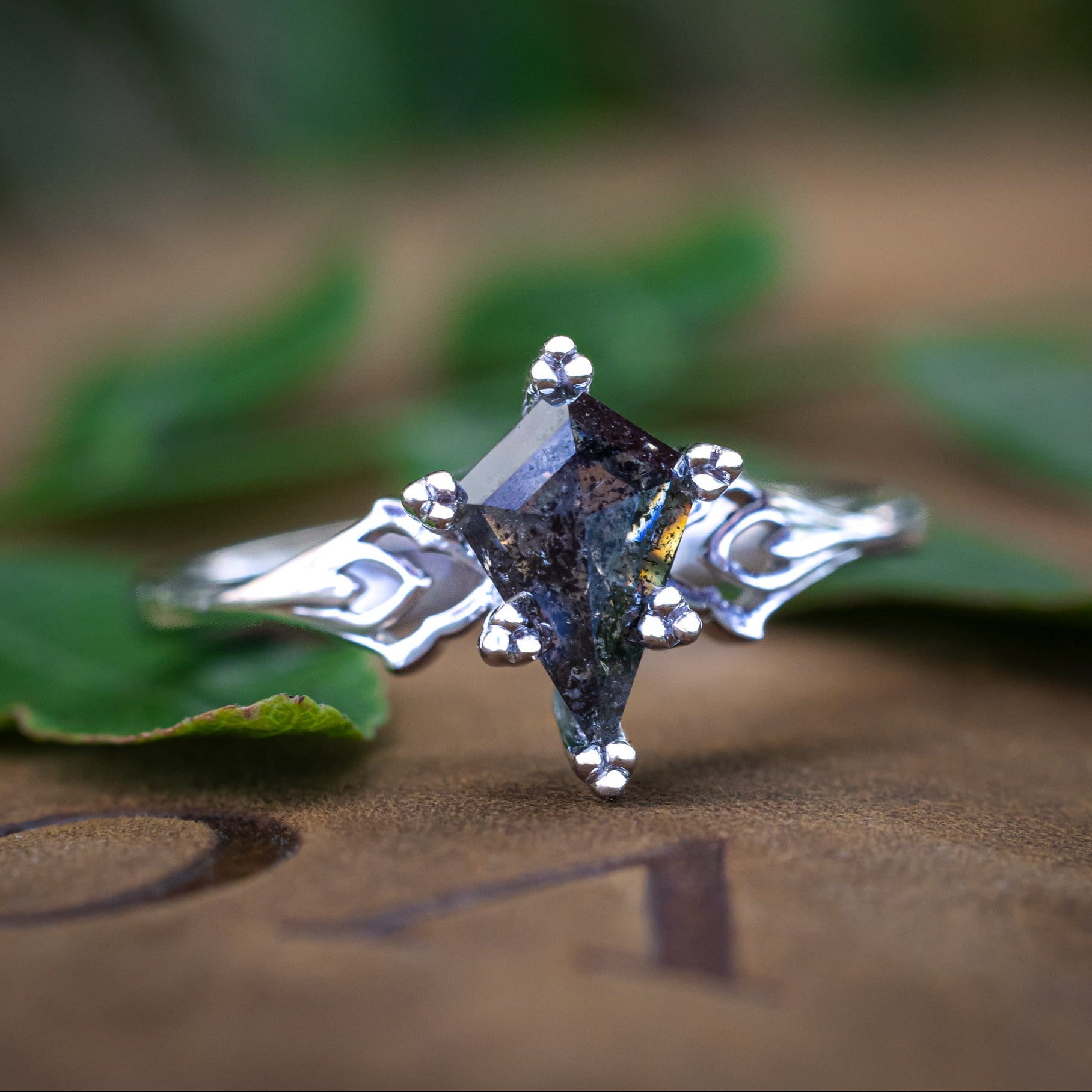 White gold ring with a kite cut salt & pepper diamond on a textured surface with green leaves in the background