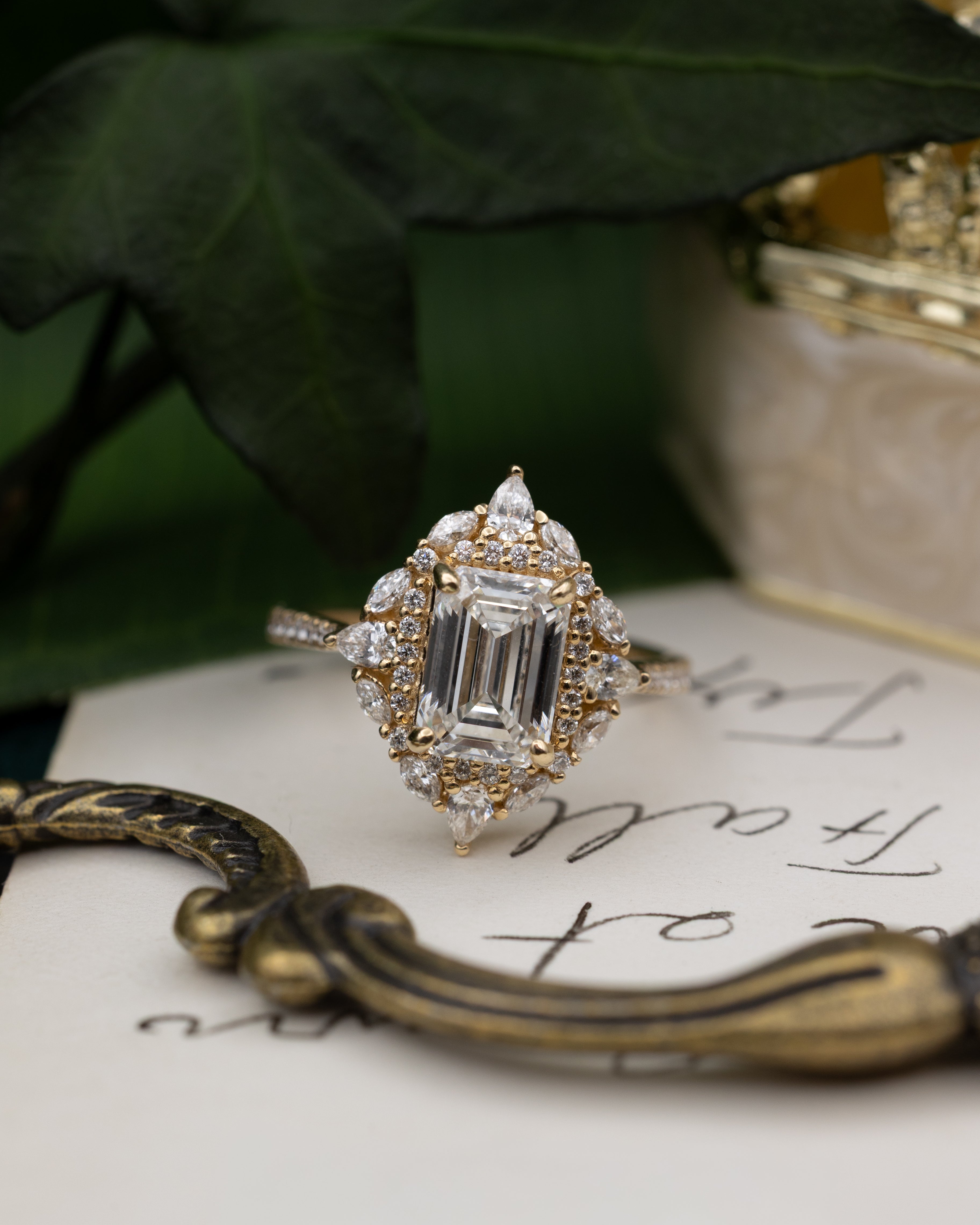 Diamond ring with emerald-cut center stone on a piece of paper with writing, surrounded by green leaves.