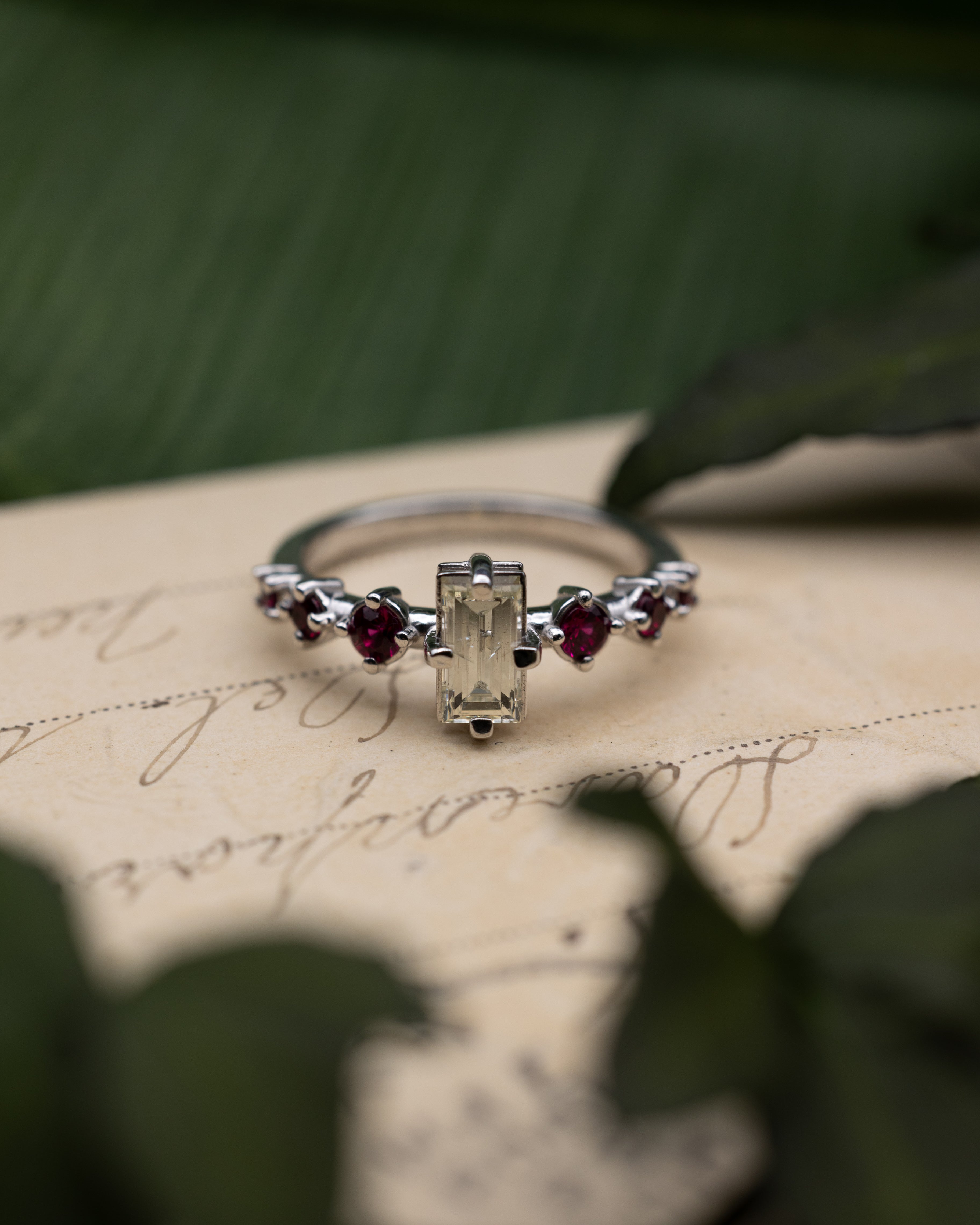 White gold ring with a champagne diamond center stone and ruby accents on a vintage letter against a blurred green background