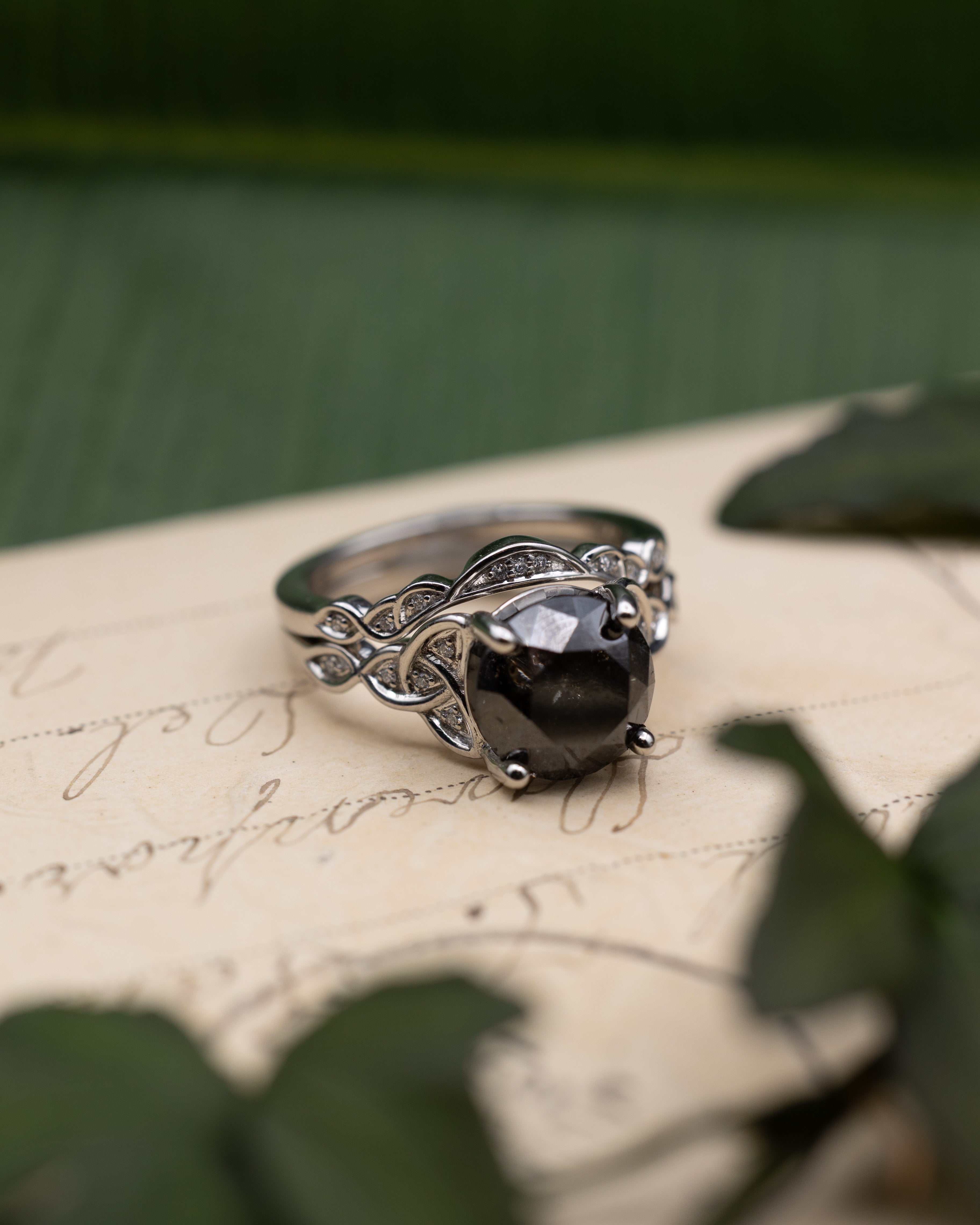 White gold ring with salt & pepper diamond center stone with diamond accents and tracer on a vintage letter against a blurred green background
