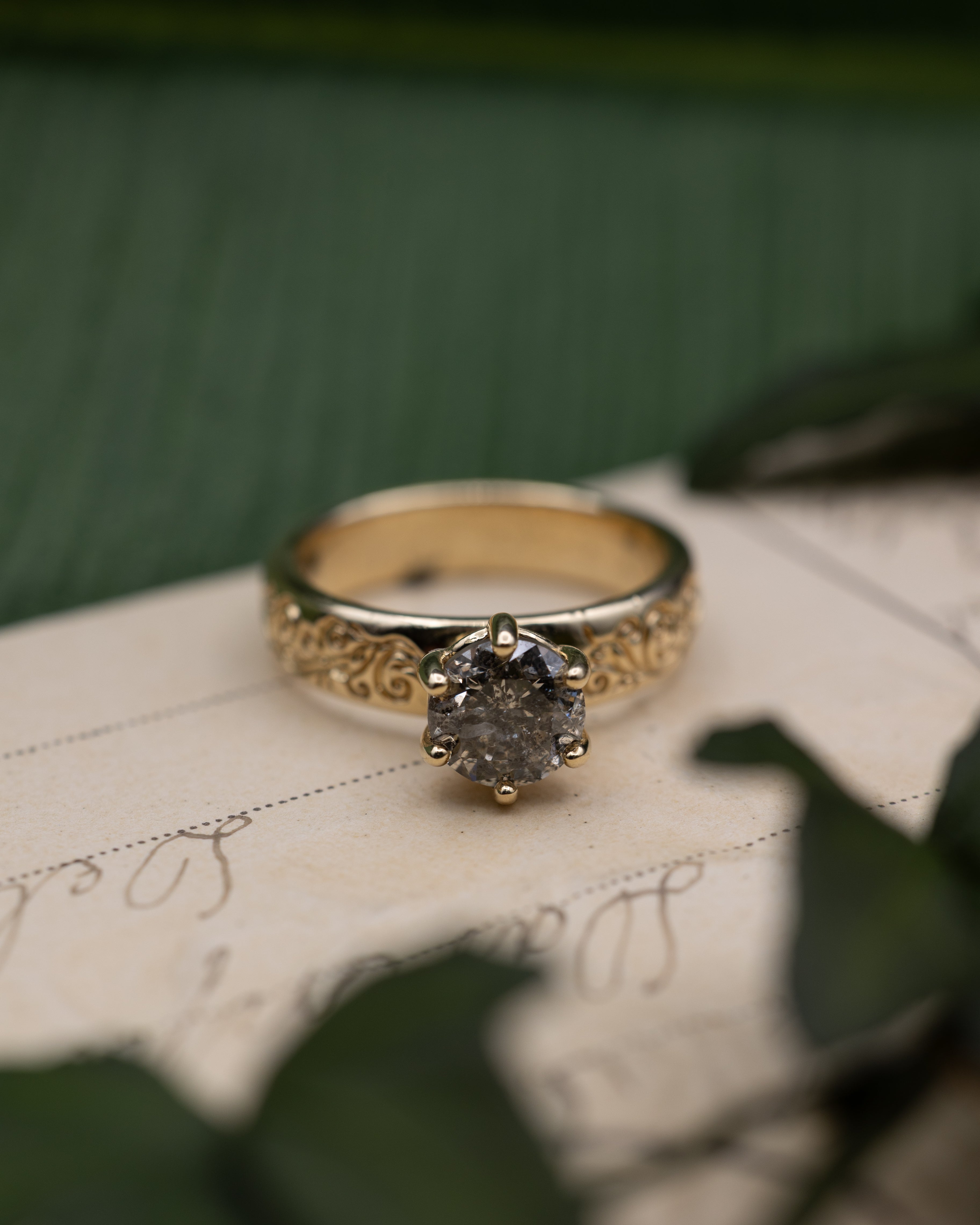 Yellow gold ring with salt & pepper diamond center stone on a vintage letter against a blurred green background