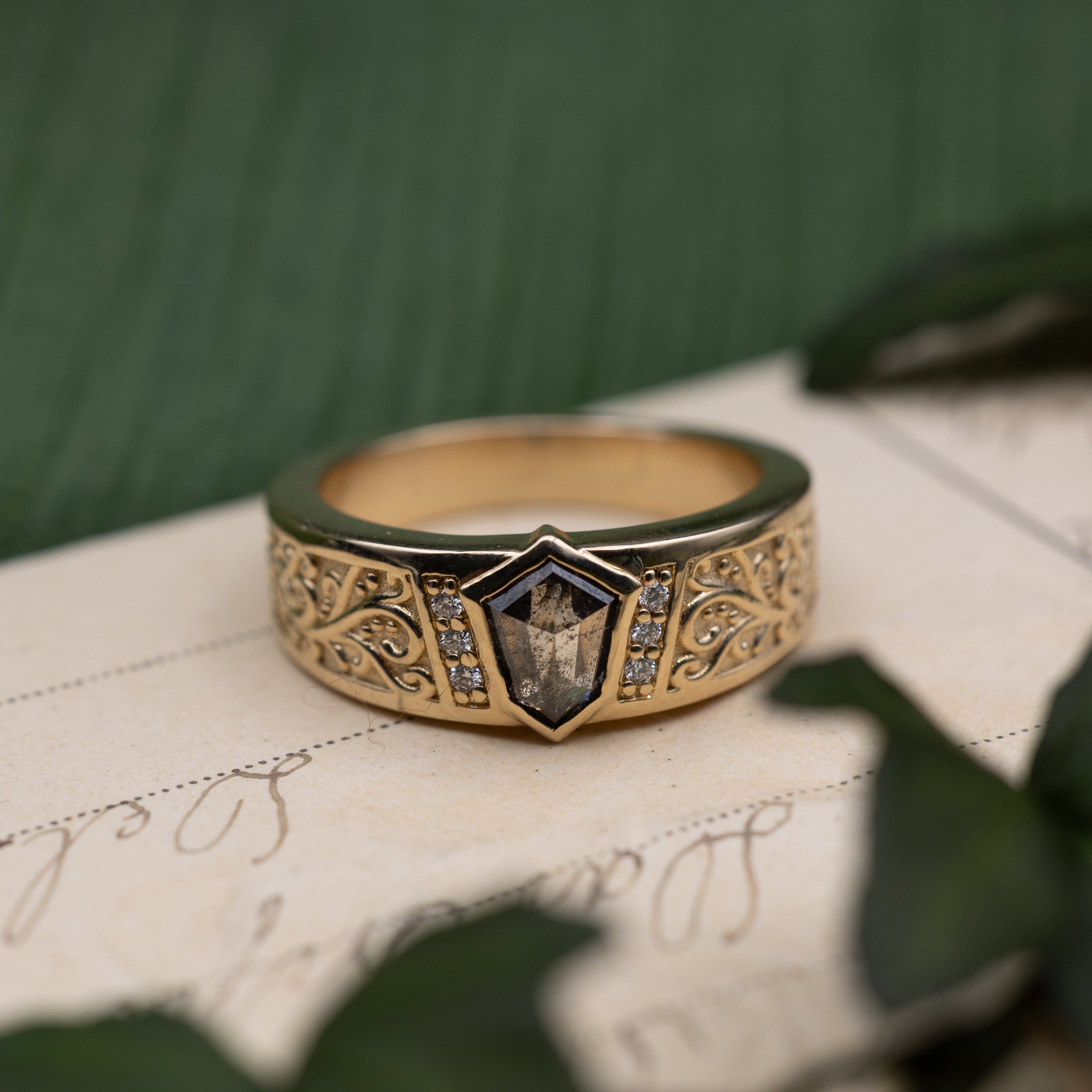 Yellow gold band with salt & pepper diamond center stone and diamond accents on a vintage letter against a blurred green background
