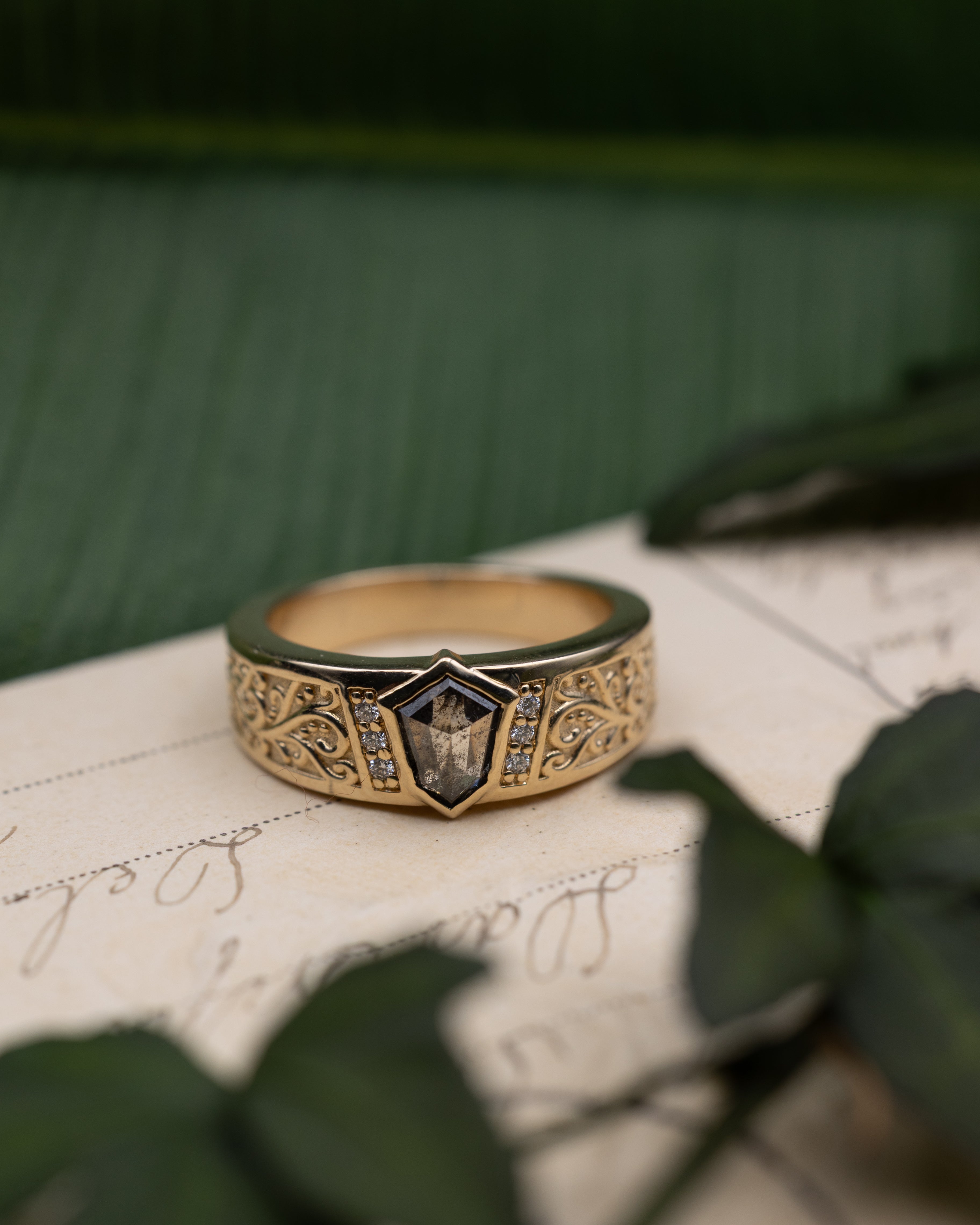 Yellow gold band with salt & pepper diamond center stone and diamond accents on a vintage letter against a blurred green background