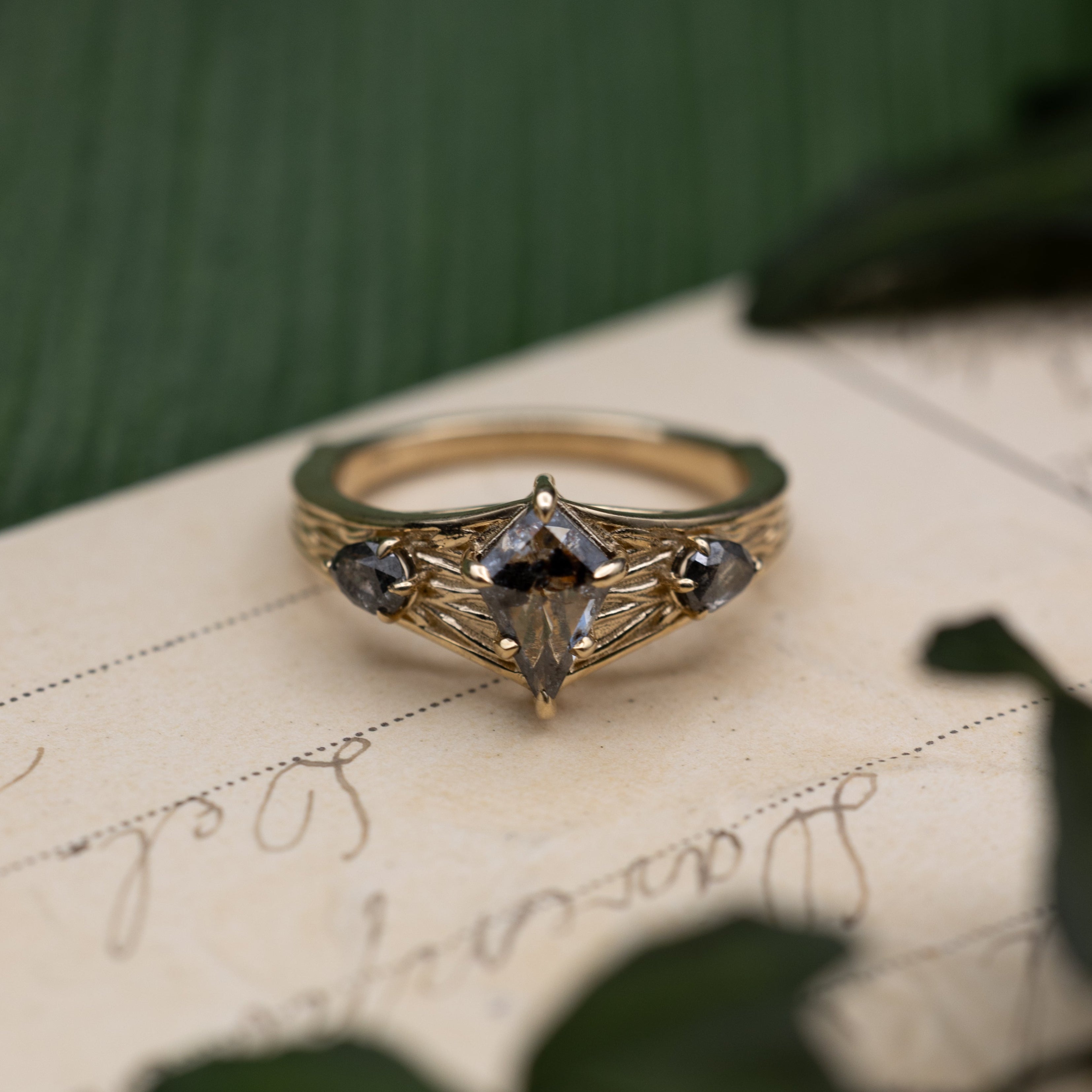 Yellow gold ring with salt & pepper diamond center stone and salt & pepper diamond accents on a vintage letter against a blurred green background