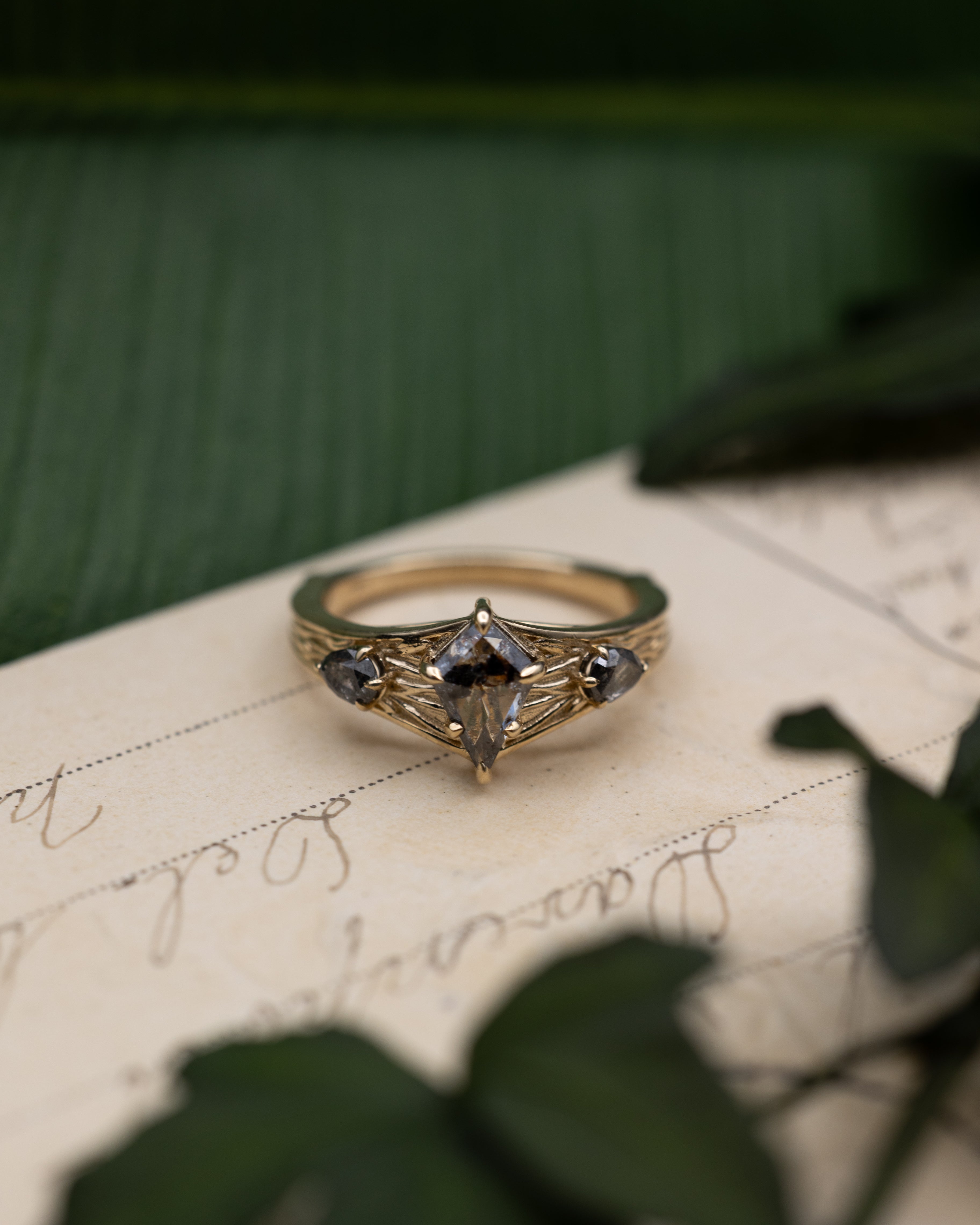 Yellow gold ring with salt & pepper diamond center stone and salt & pepper diamond accents on a vintage letter against a blurred green background
