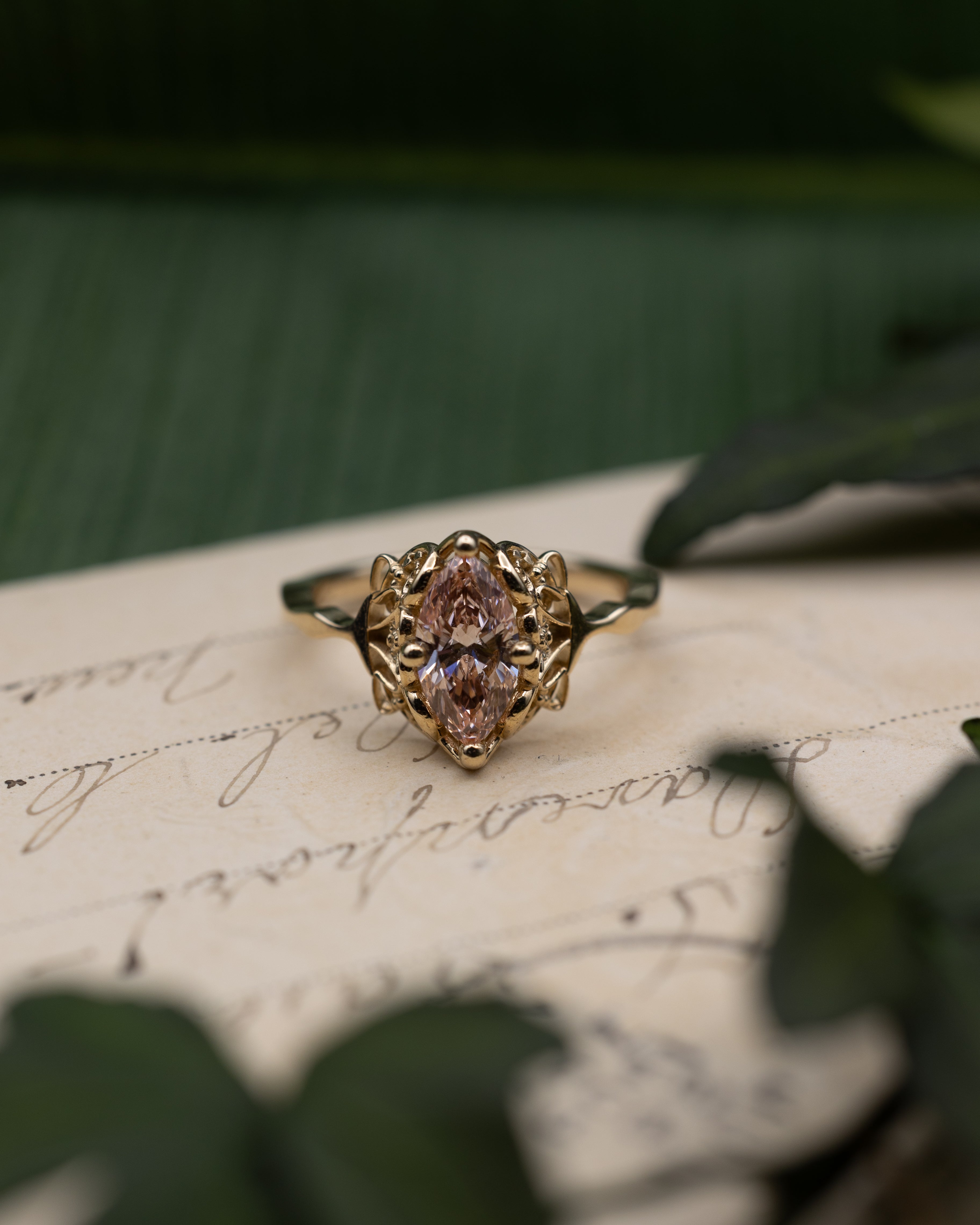 Sculptural yellow gold ring with pink diamond center stone on a vintage letter against a blurred green background