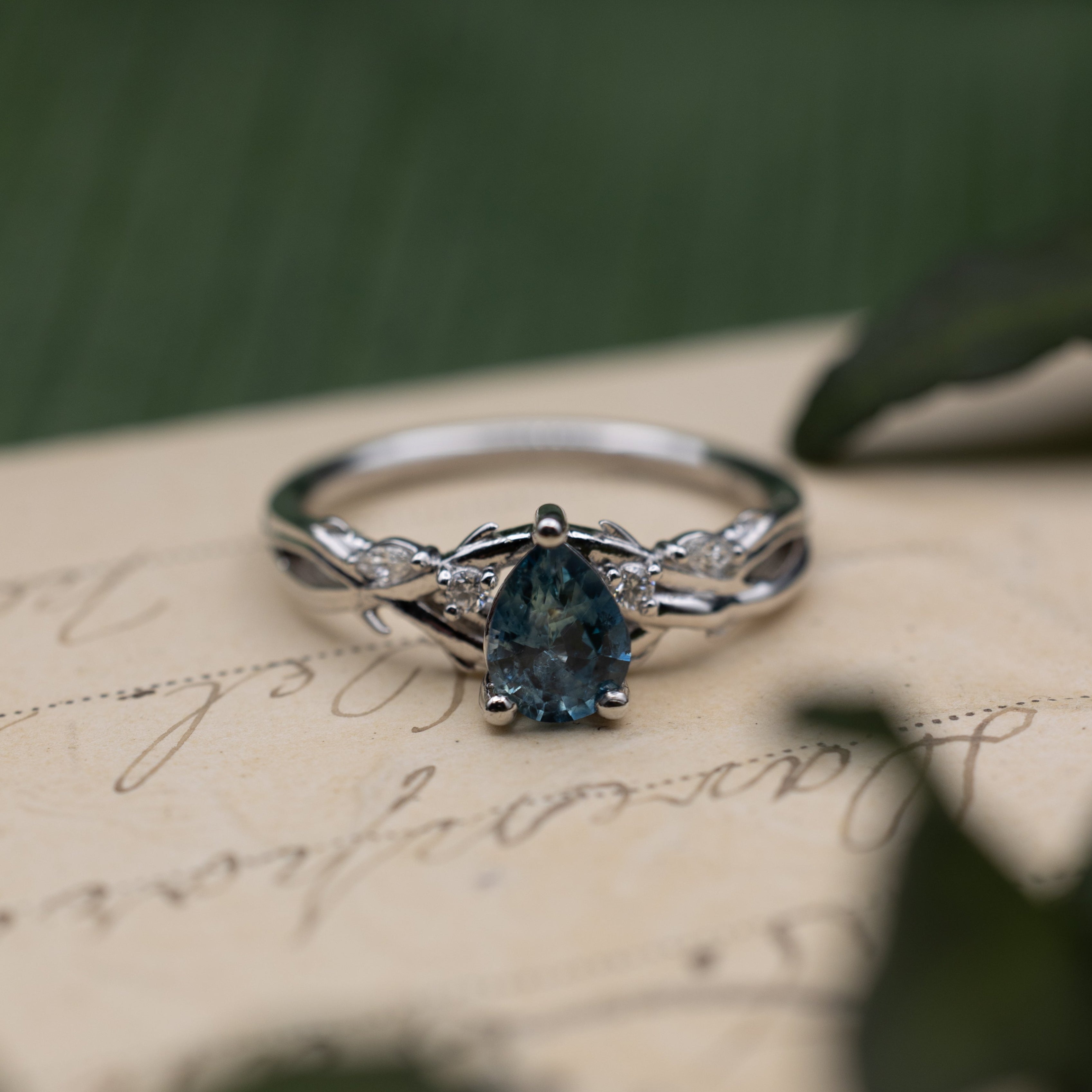 White gold ring with blue sapphire center stone and diamond accents on a vintage letter against a blurred green background
