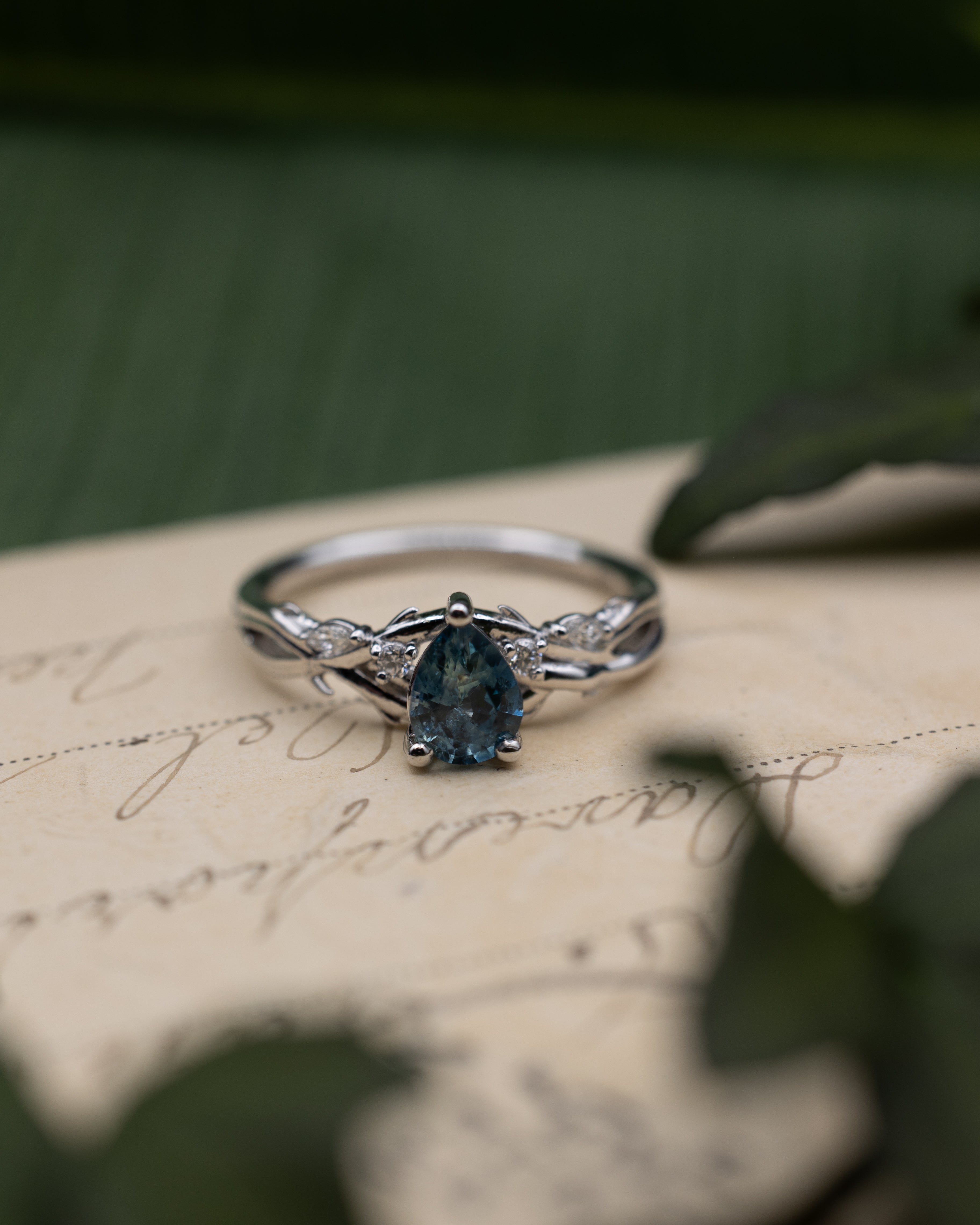 White gold ring with blue sapphire center stone and diamond accents on a vintage letter against a blurred green background