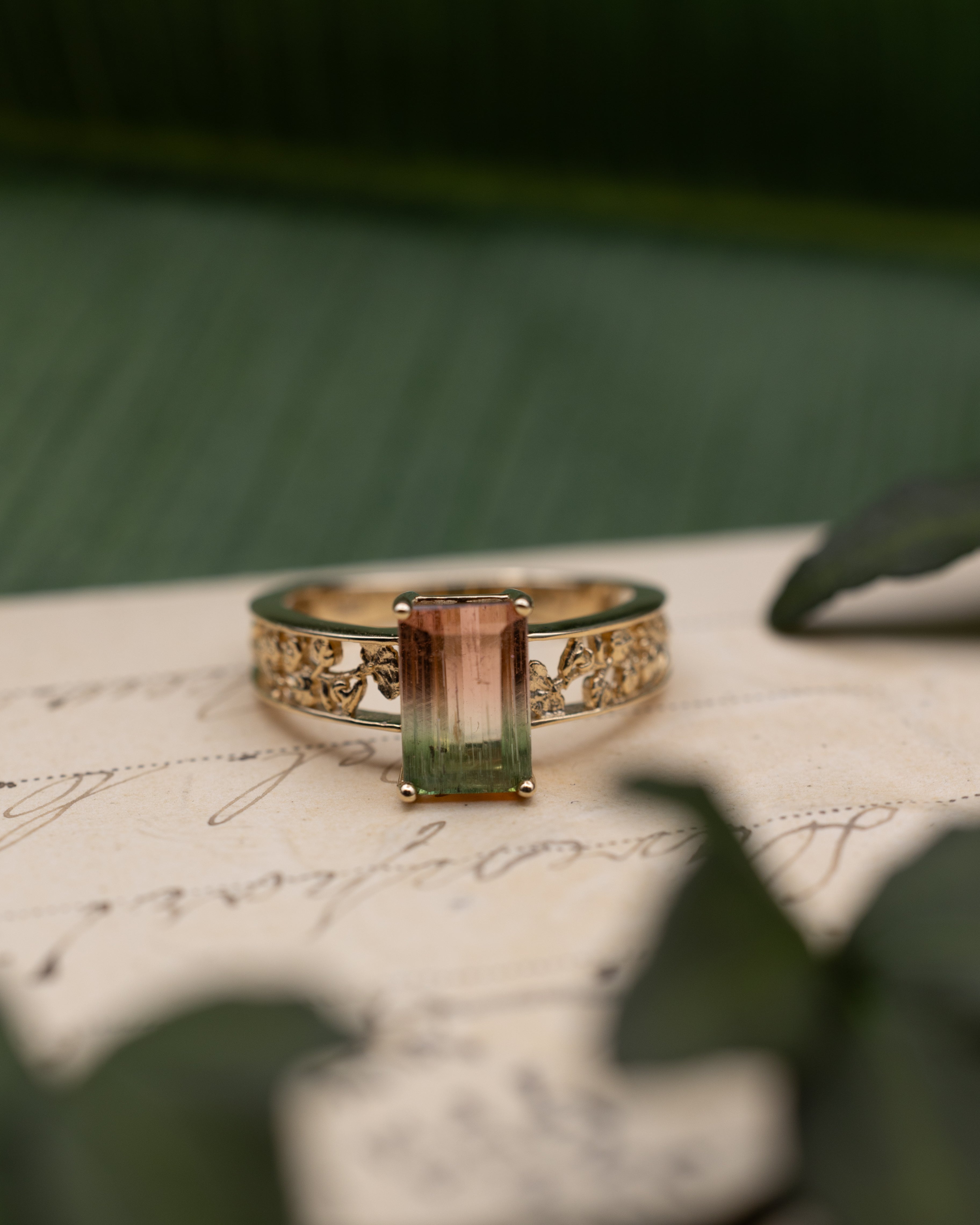 Yellow gold ring with watermelon tourmaline center stone on a vintage letter against a blurred green background