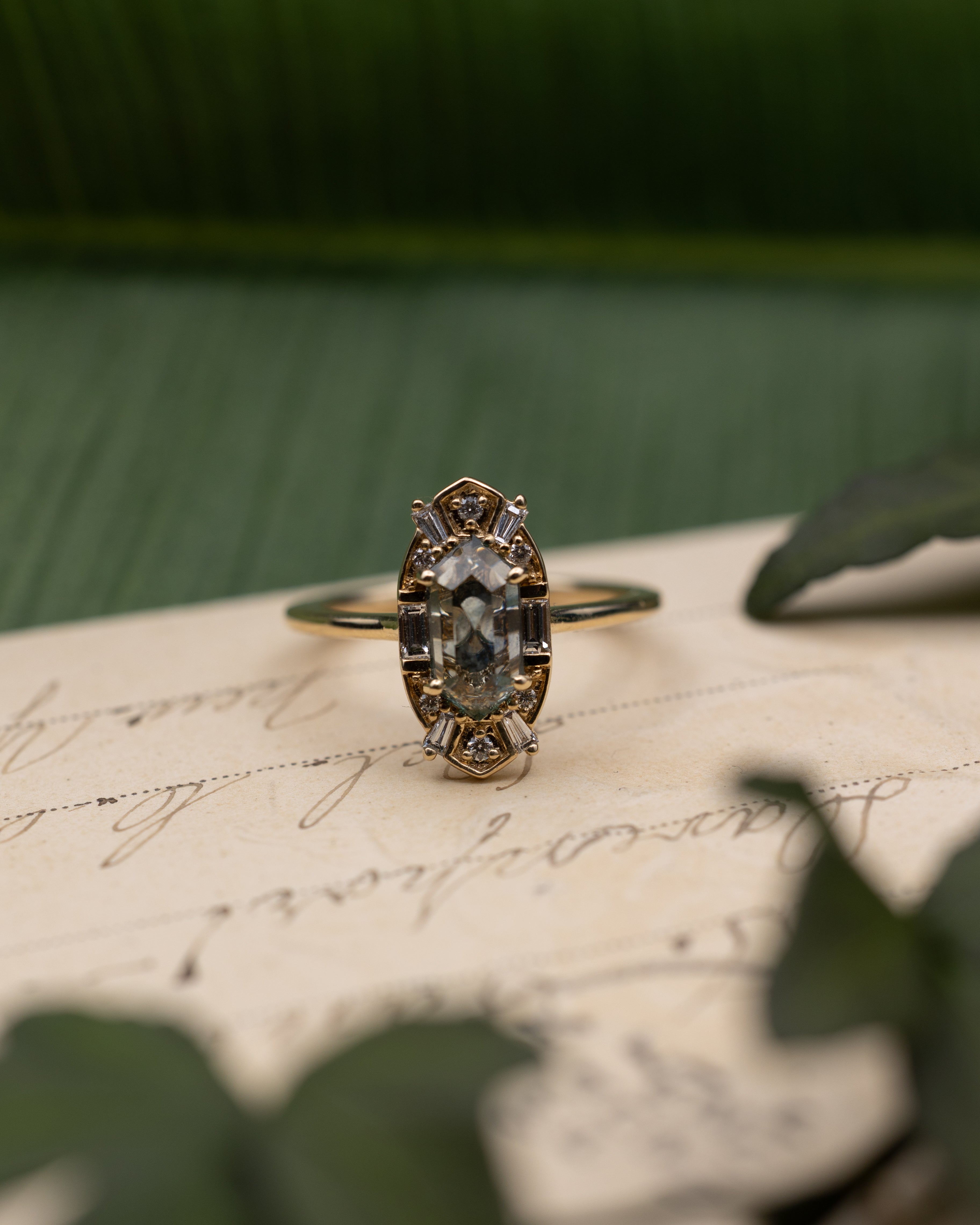 Yellow gold ring with sapphire center stone and diamond halo on a vintage letter against a blurred green background