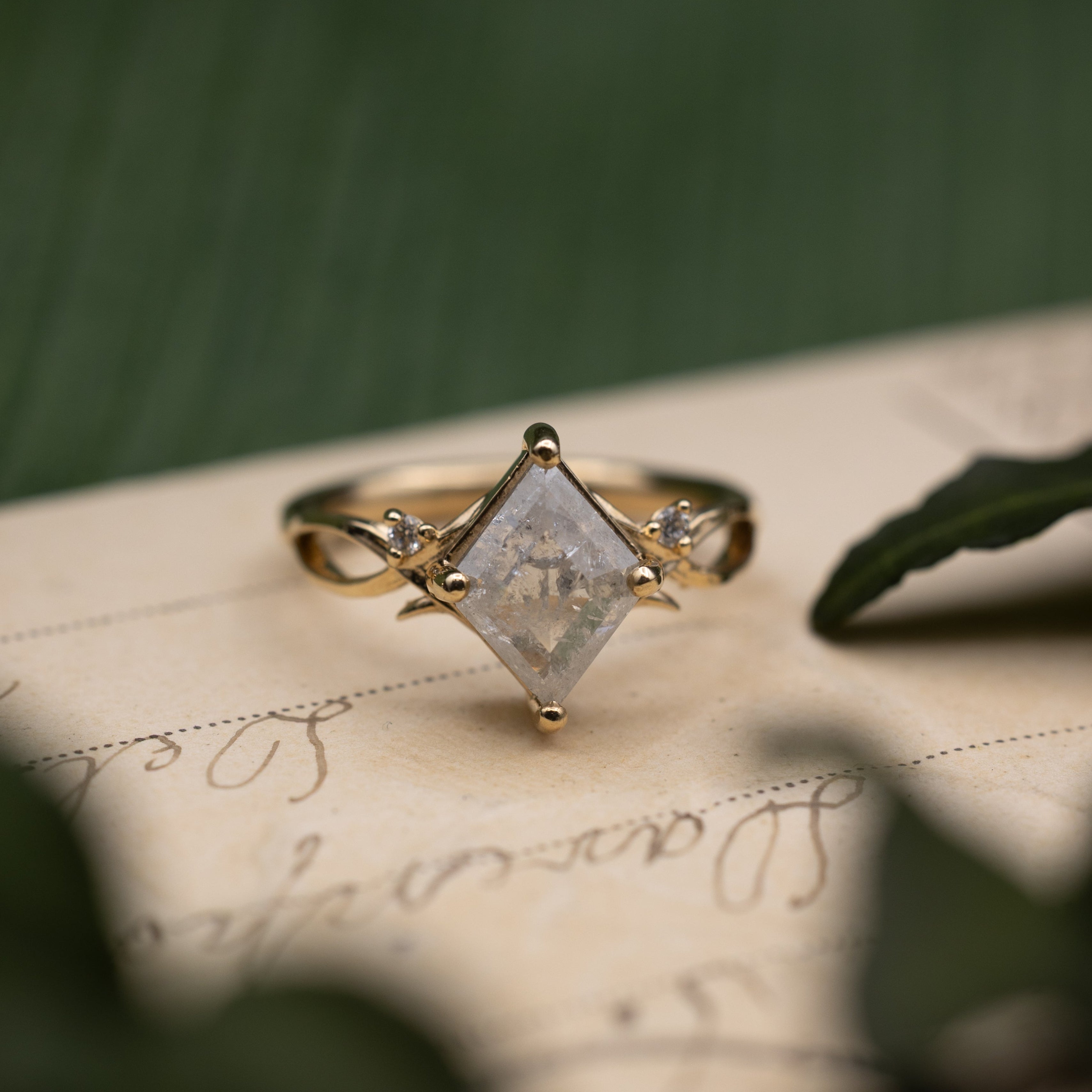 Yellow gold ring with salt & pepper diamond center stone sitting on a vintage letter against a blurred green background