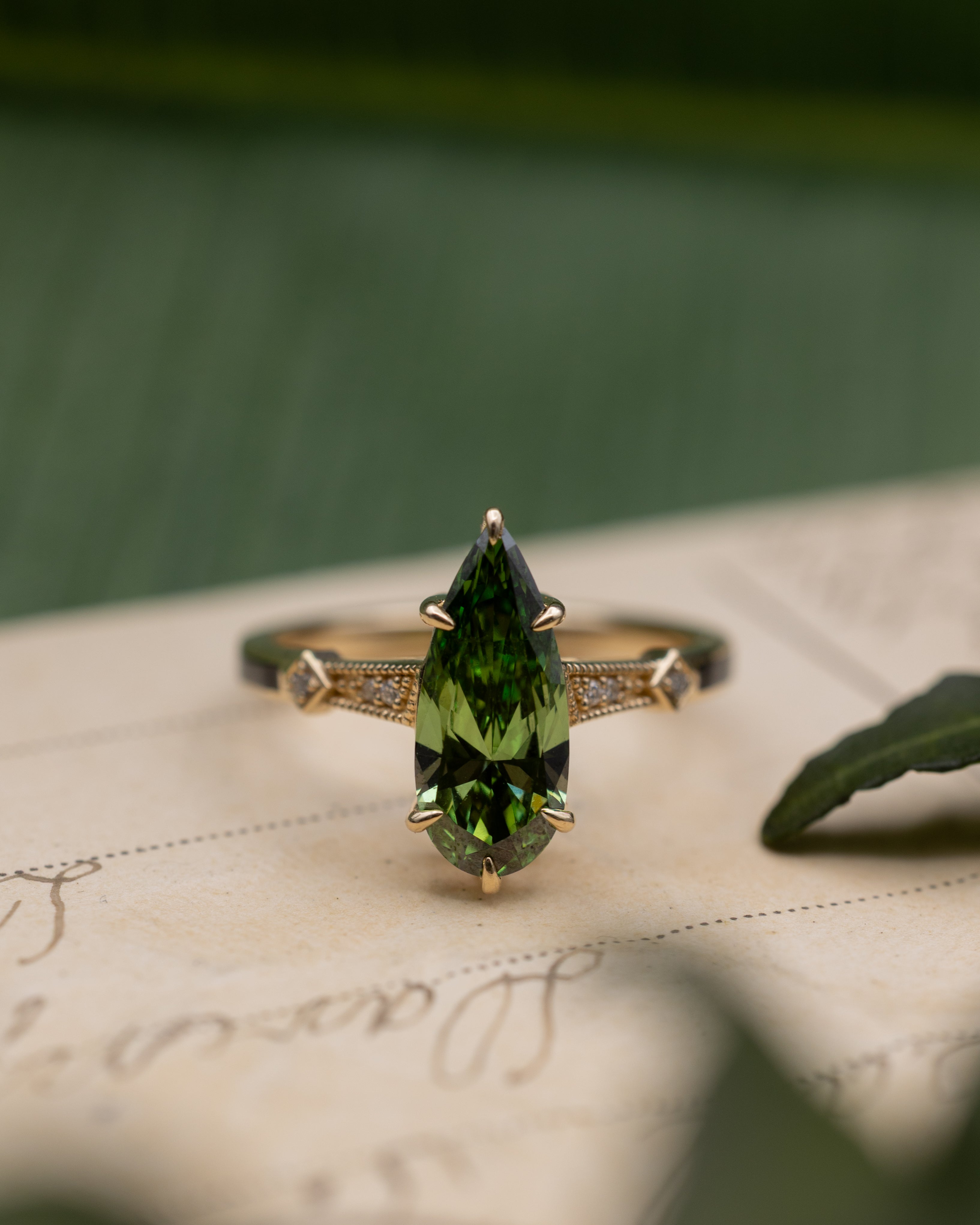Sculptural yellow gold ring with green diamond center stone sitting on a vintage letter against a blurred green background