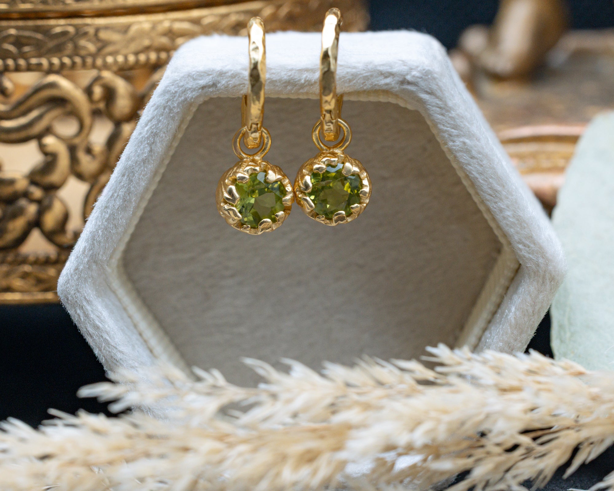 Gold earrings with green peridot on a white hexagonal stand.