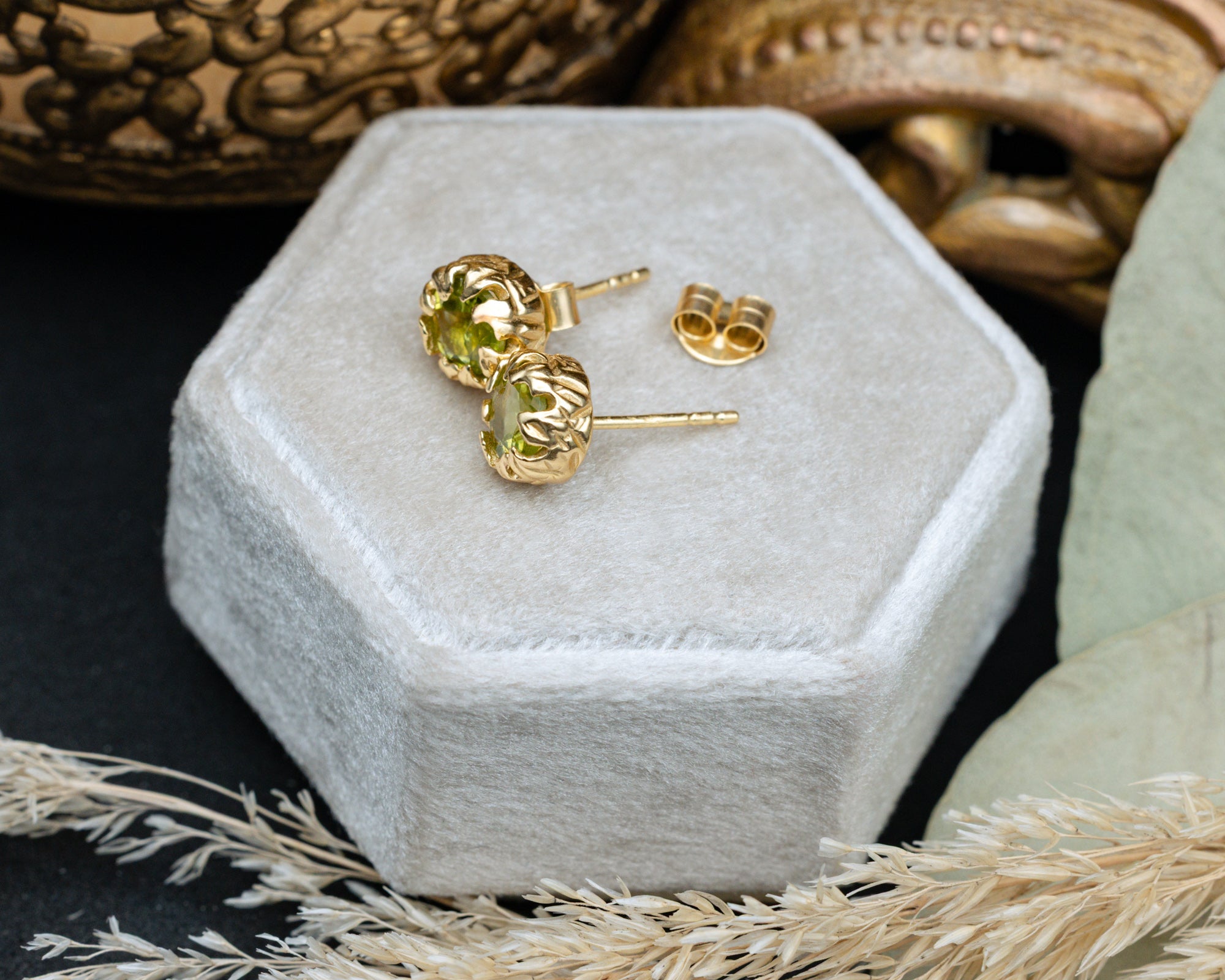 Gold peridot earrings on a hexagonal gray velvet box with natural elements around.