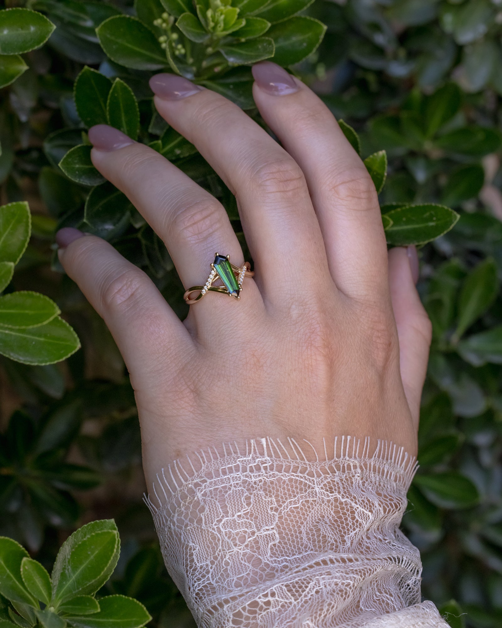 Hand wearing gold ring with a green tourmaline and diamond accents on a leafy background