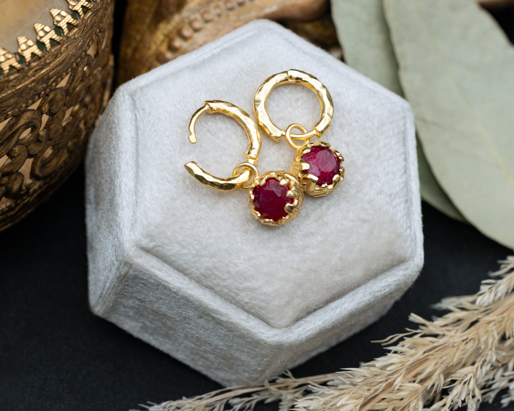 Gold hoop earrings with red rubies on a white hexagonal stand.