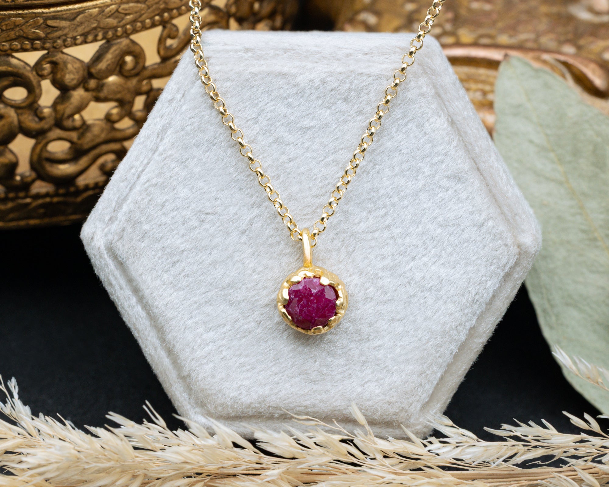 Gold necklace with a red ruby pendant on a white cushion, with a decorative background.