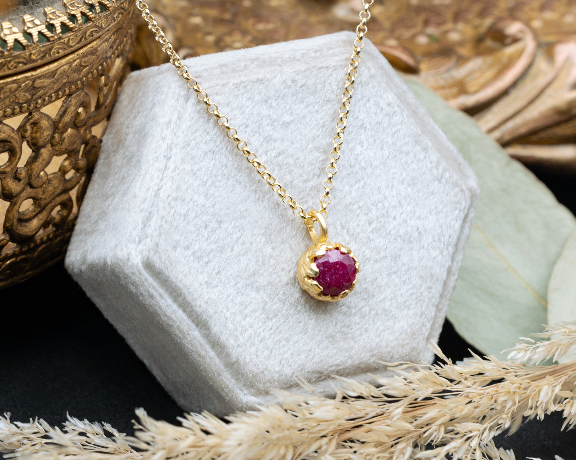 Gold necklace with a red ruby pendant on a white hexagonal stand.