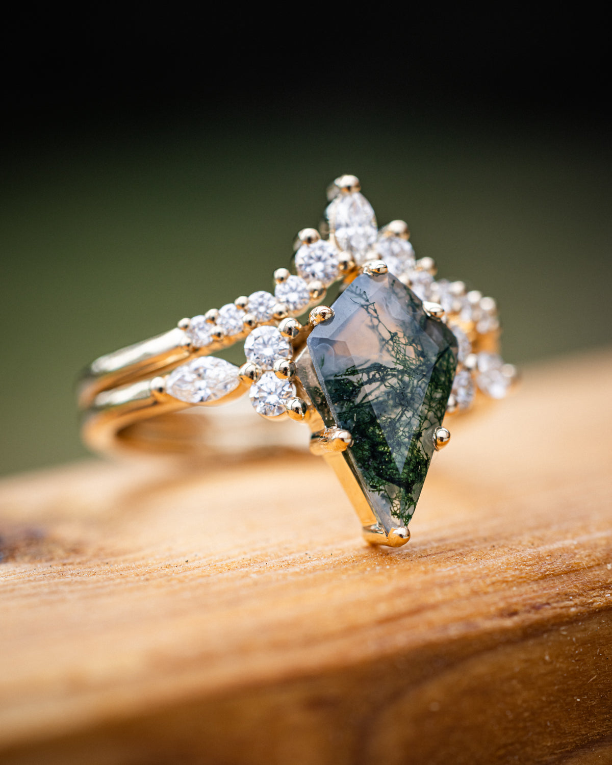 Gold ring with moss agate center stone and diamond accents on a wooden surface and green background