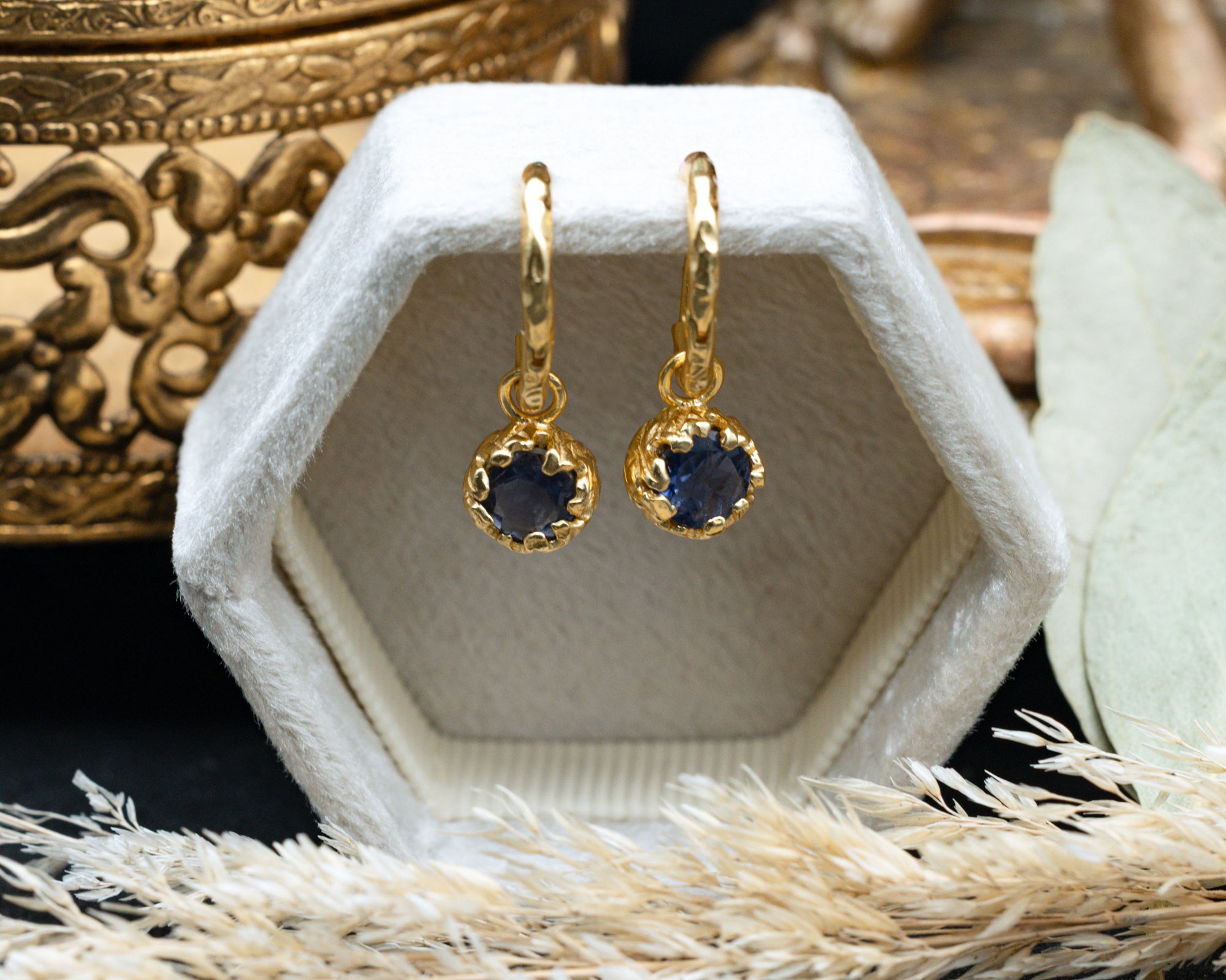 Gold earrings with blue sapphires in a white hexagonal jewelry box on a decorative surface.