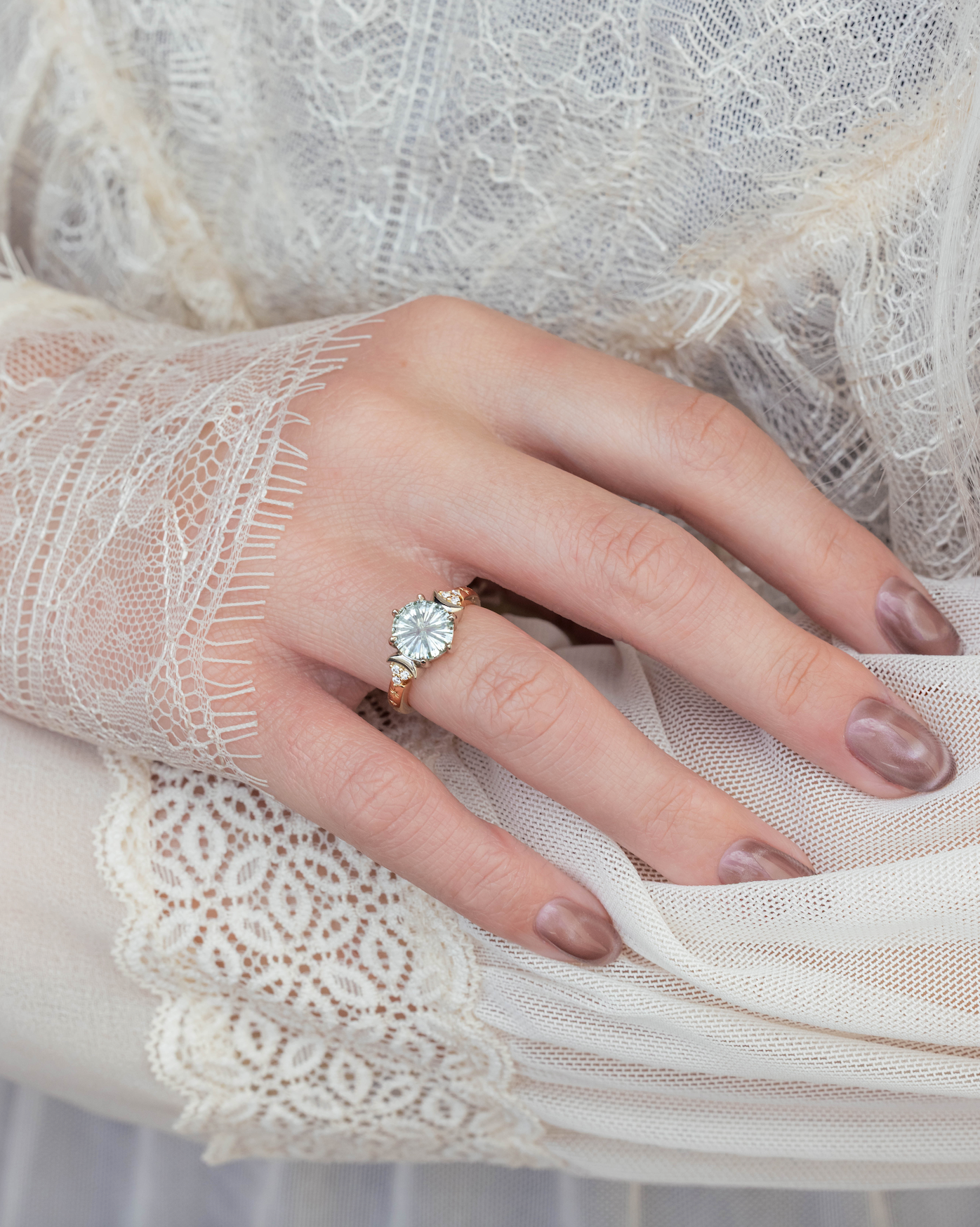 Close-up of a hand wearing a green sapphire engagement ring with a white lace sleeve.