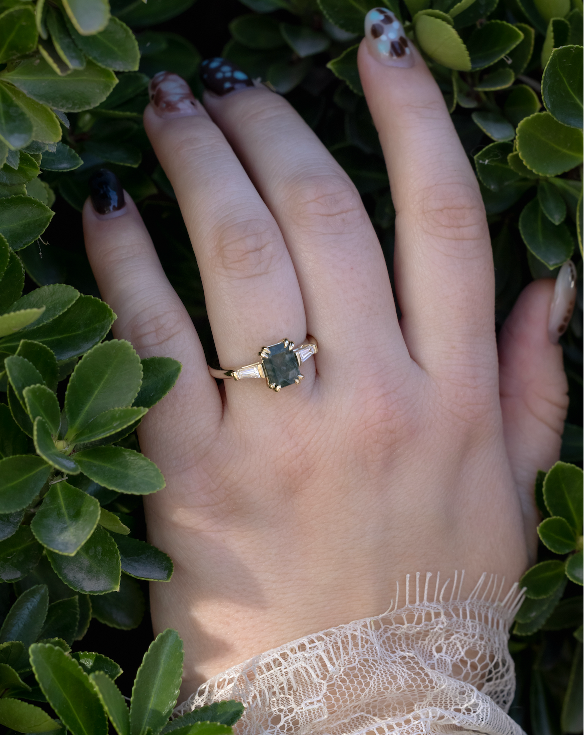 Lace sleeved hand wearing gold ring with moss agate center stone & diamond accents against a greenery background