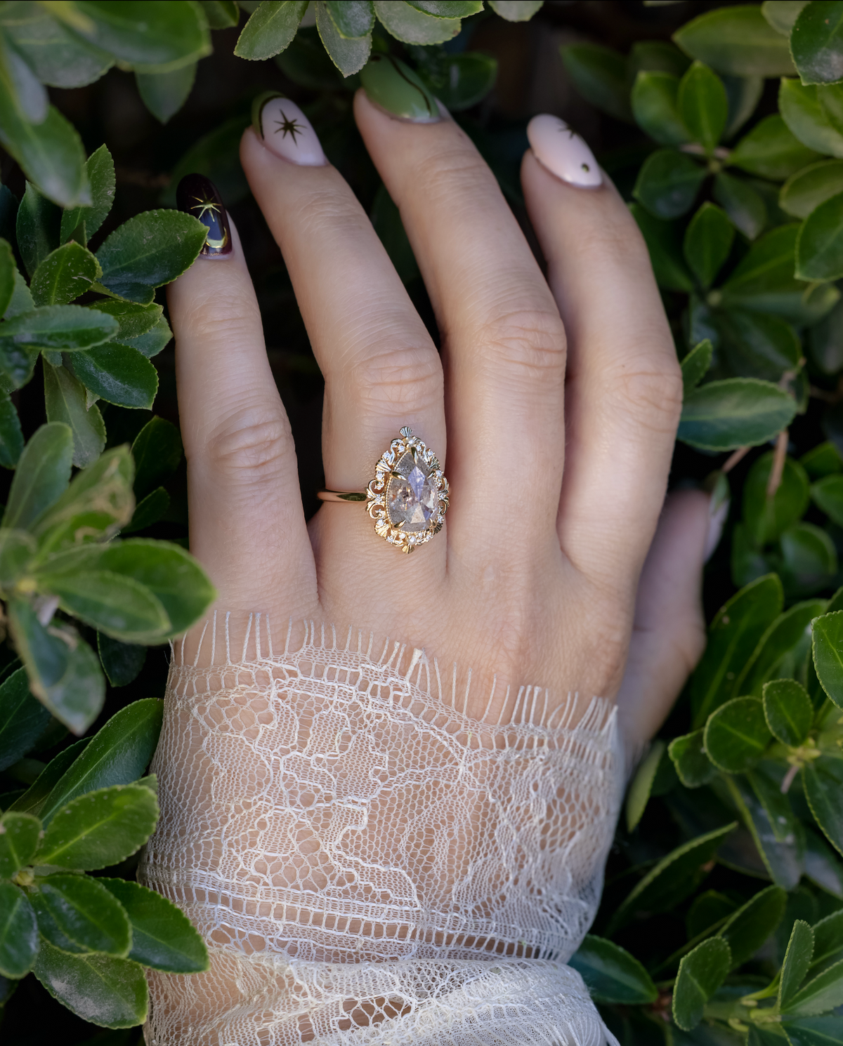 Hand wearing gold ring with salt & pepper diamond with lace sleeve against greenery