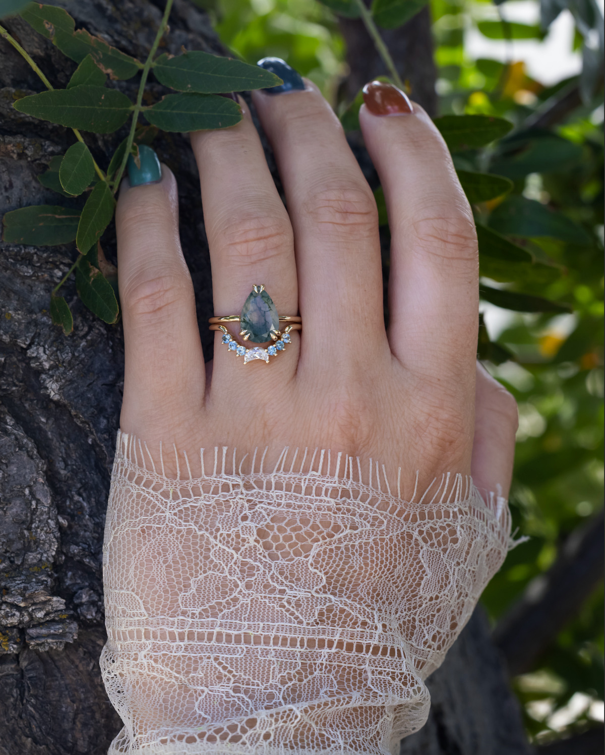 Hand wearing gold moss agate engagement ring featuring tracer with moissanite and Montana sapphire accents against a green tree