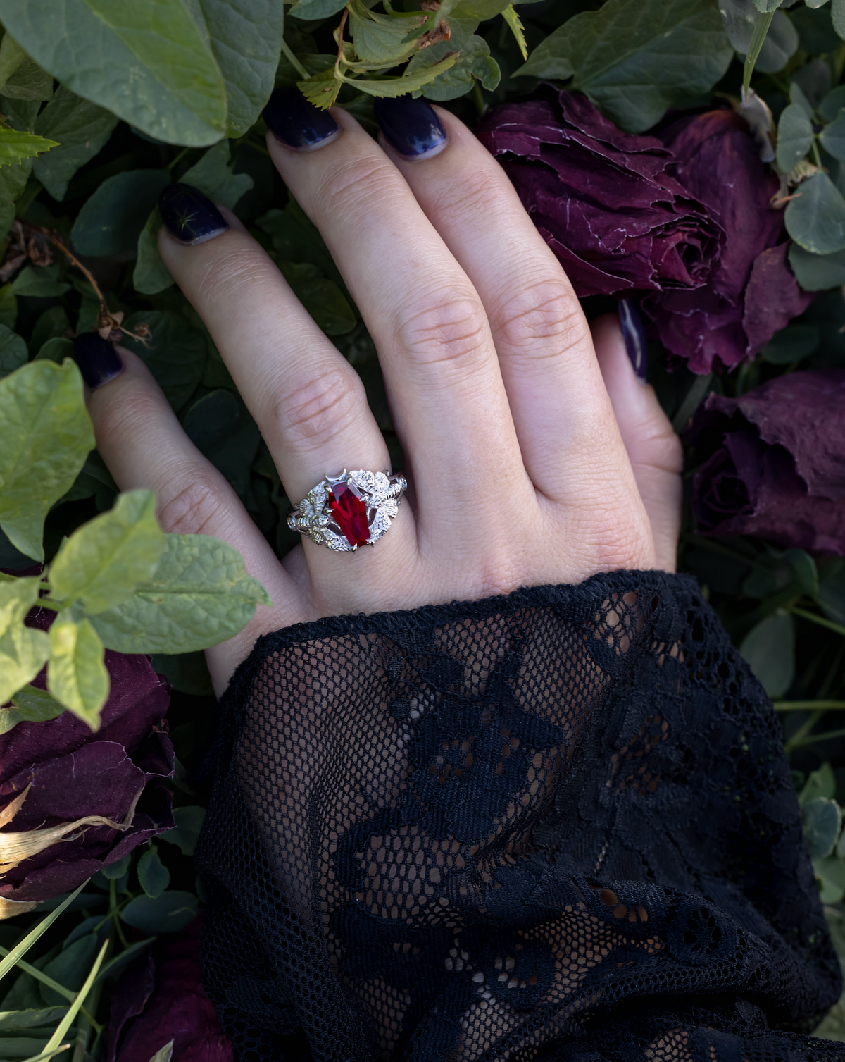 Lace sleeved hand wearing white gold engagement ring with deadhead moth filigree, coffin cut ruby center stone, and diamond accents against a rose bush
