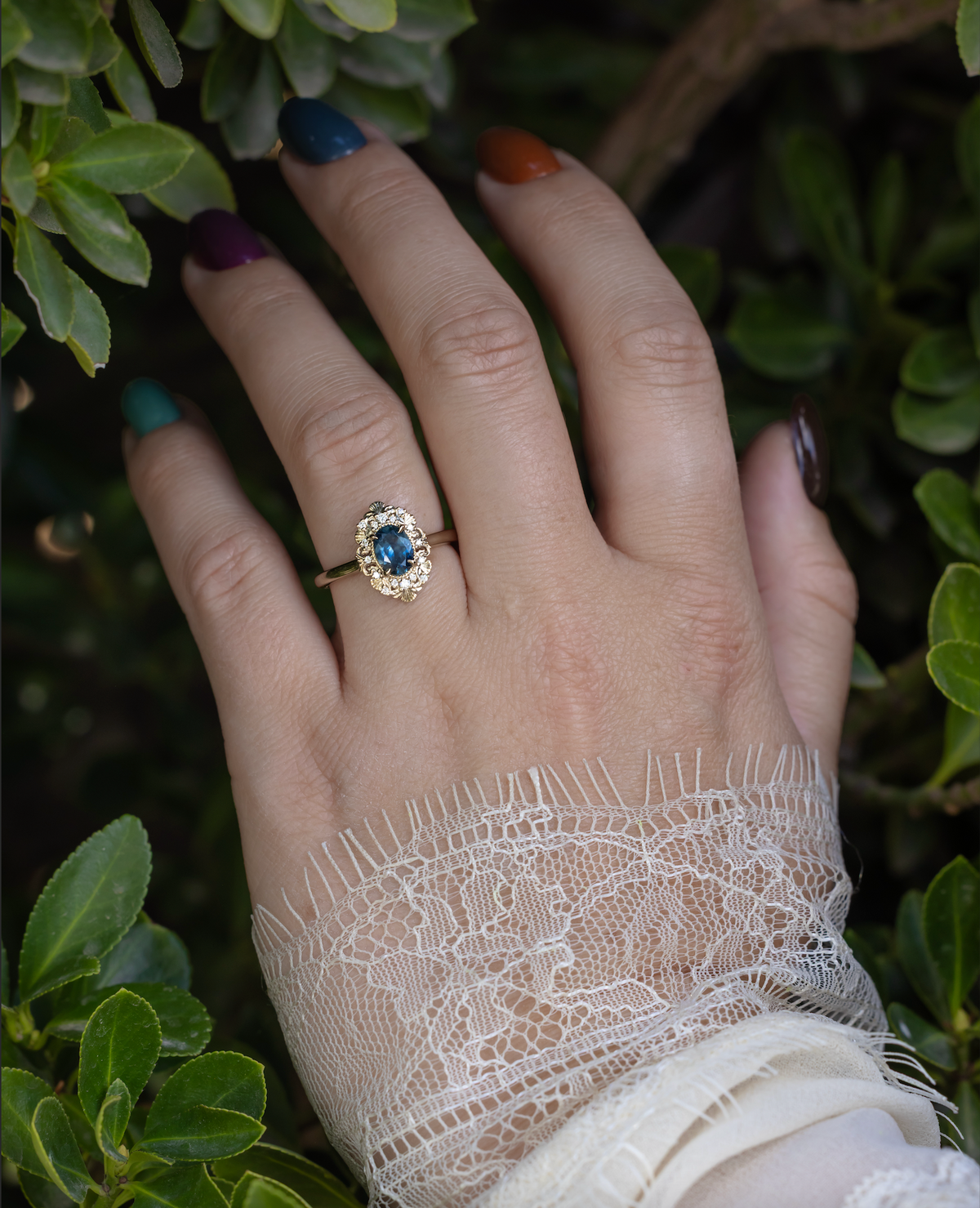 Hand wearing a gold ring with Montana sapphire and diamond accents against a green foliage background