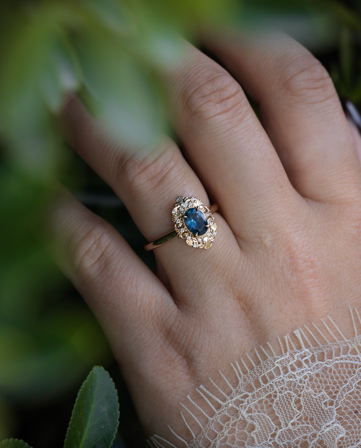 Hand wearing a gold ring with Montana sapphire and diamond accents against a green foliage background