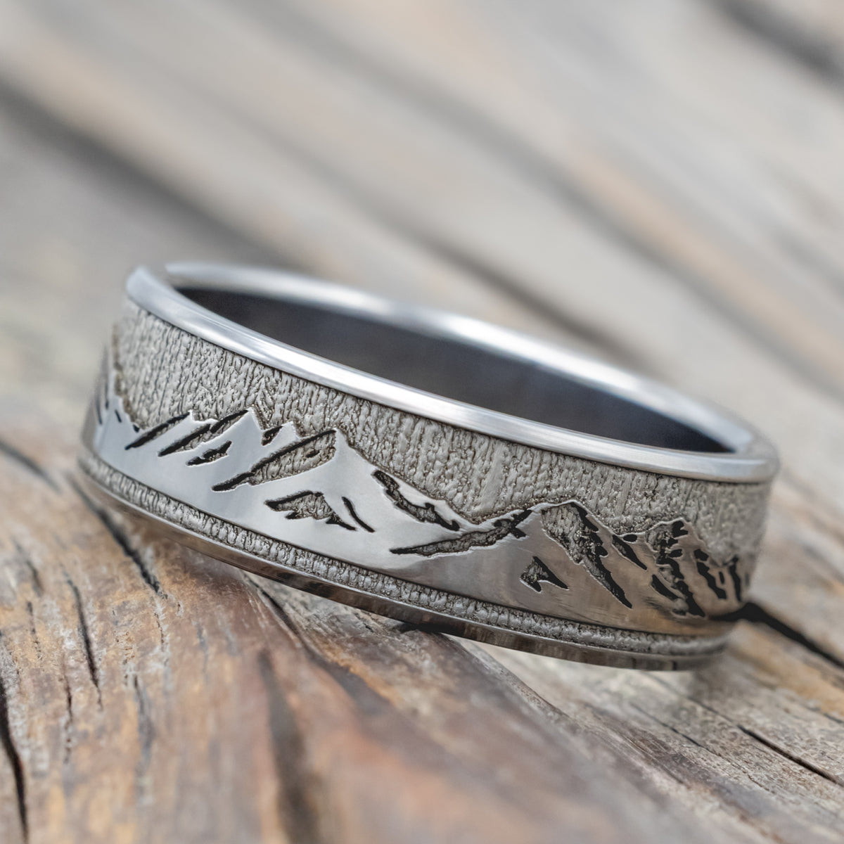 A titanium mountain ring with an engraved landscape design, resting on a rustic wooden surface.