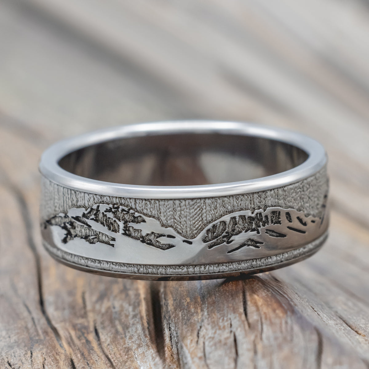 A titanium mountain ring with an engraved landscape design, resting on a rustic wooden surface.
