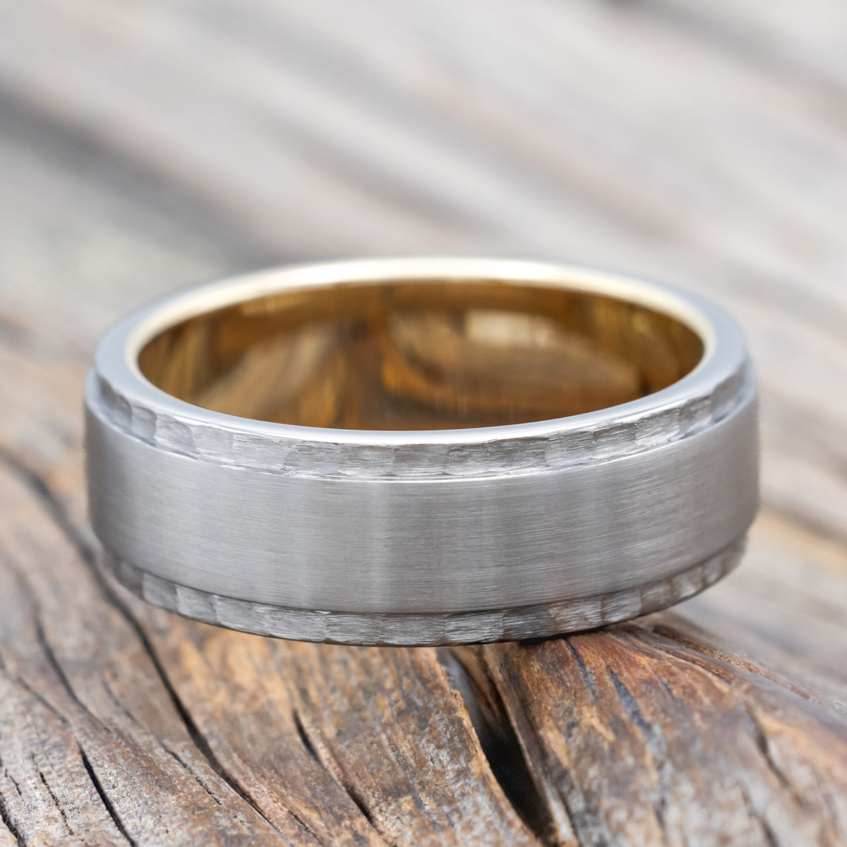 A titanium wedding ring with brushed center and hammered edges with yellow gold lining on a wooden surface
