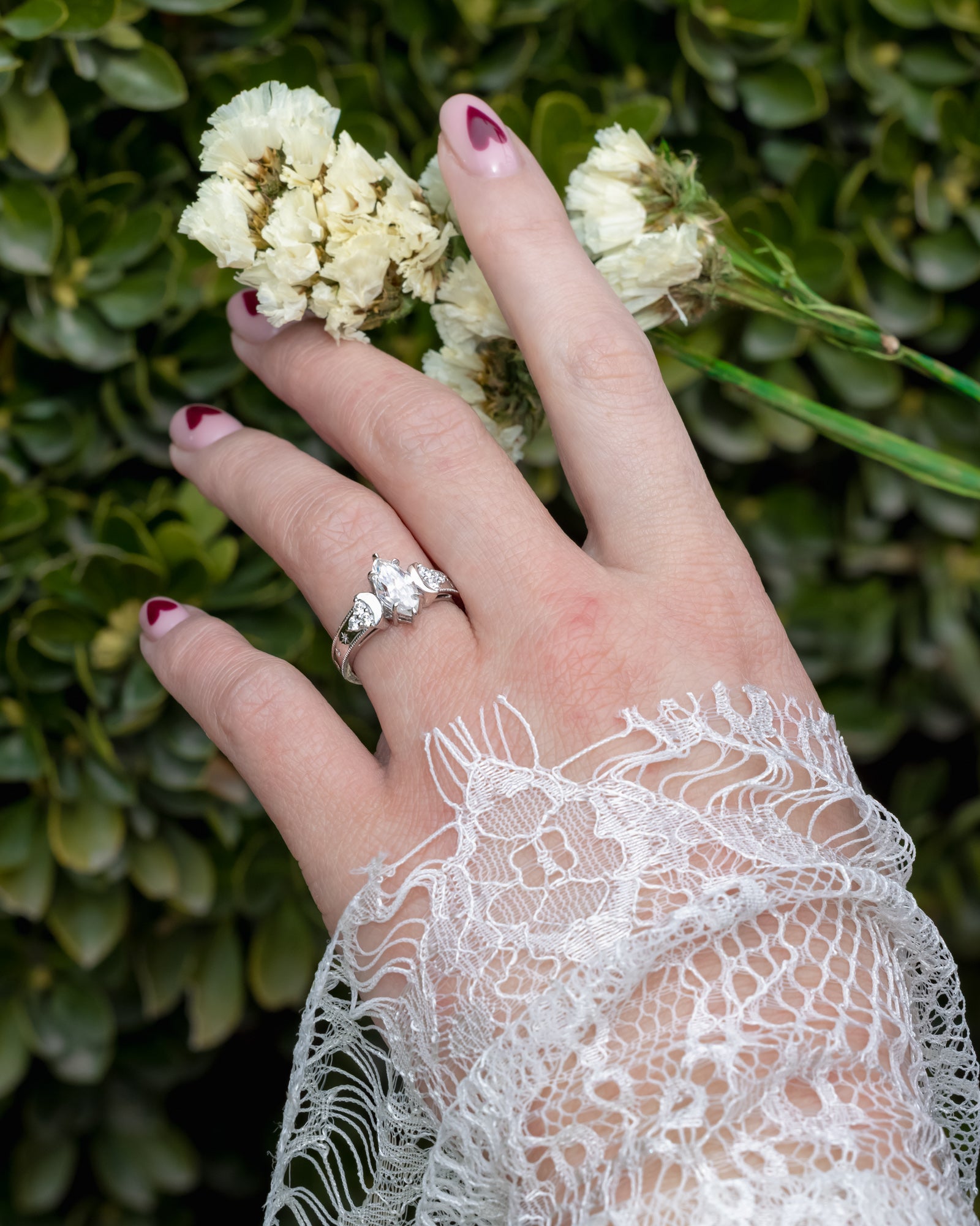 Hand wearing a moissanite ring with lace sleeve against a green leafy background
