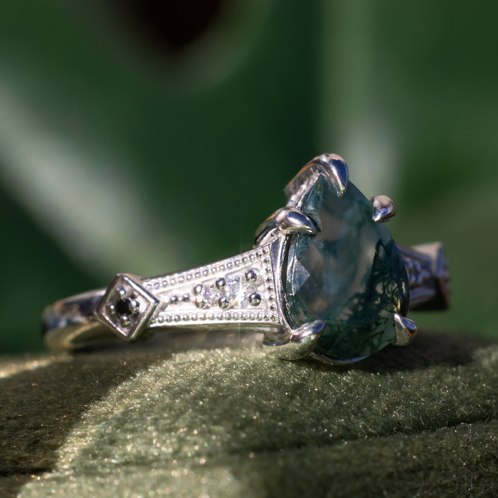 White gold engagement ring with moss agate and diamond accents on a green surface