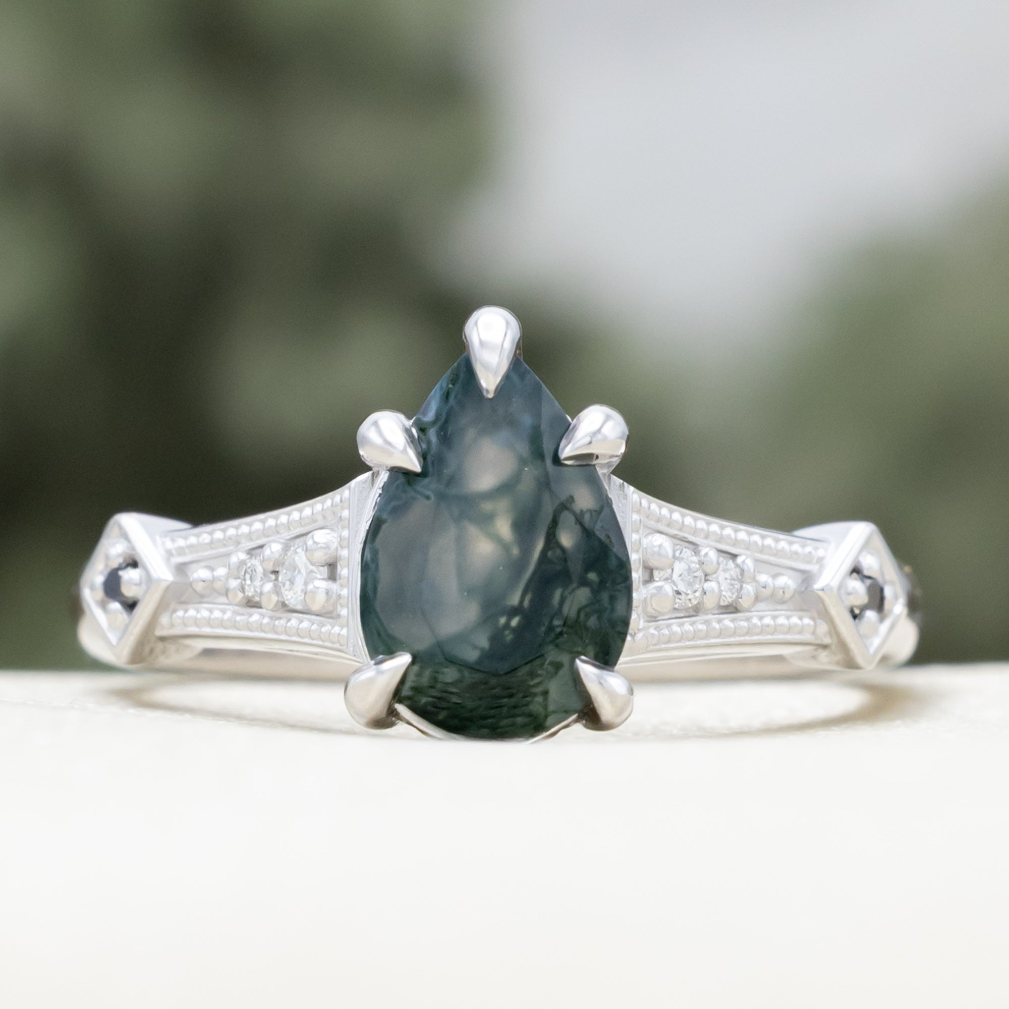 White gold engagement ring with moss agate and diamond accents on a blurred background