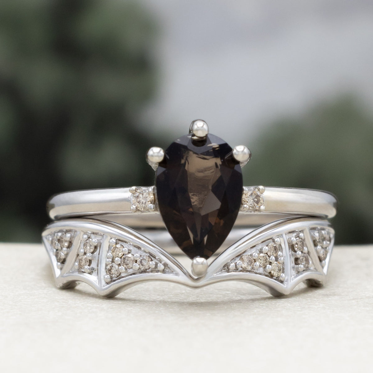White gold engagement ring with smoky quartz center stone and bat wing bottom tracer with champagne diamond accents against a blurred green background