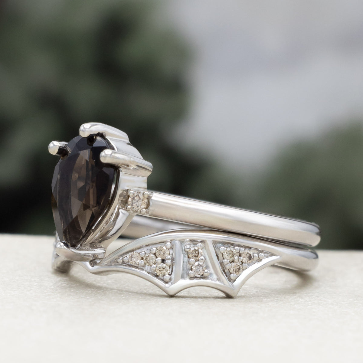 White gold engagement ring with smoky quartz center stone and bat wing bottom tracer with champagne diamond accents against a blurred green background
