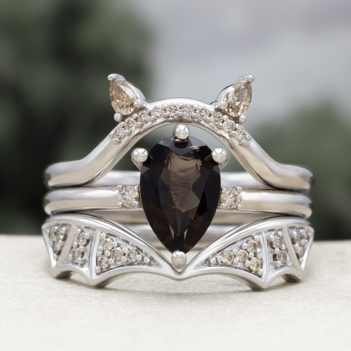White gold engagement ring with smoky quartz center stone, bat ear top tracer, and bat wing bottom tracer with champagne diamond accents against a blurred green background