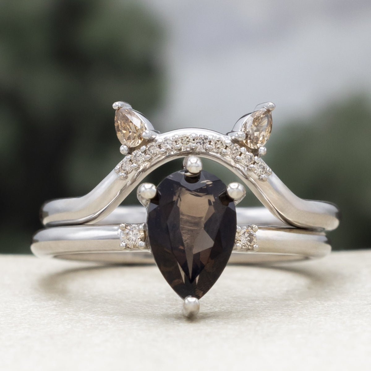 White gold engagement ring with smoky quartz center stone and bat ear top tracer with champagne diamond accents against a blurred green background