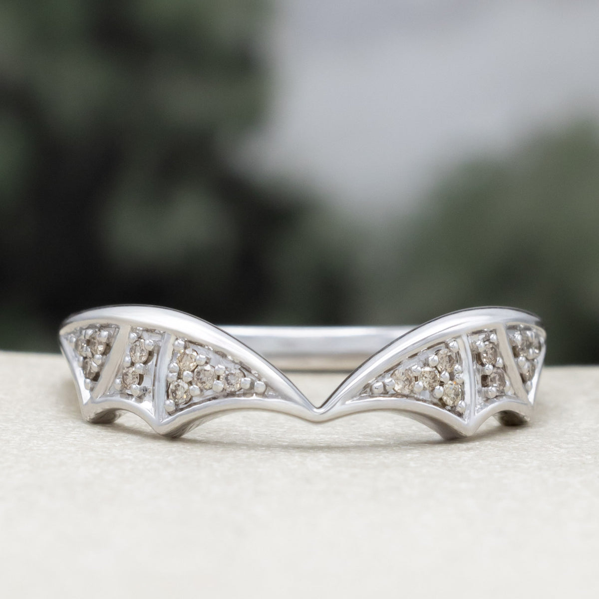 White gold bat wing wedding ring tracer with champagne diamond accents against a blurred green background
