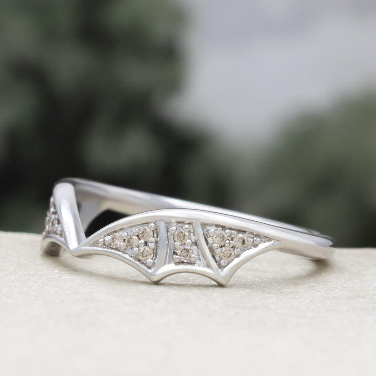 White gold bat wing wedding ring tracer with champagne diamond accents against a blurred green background