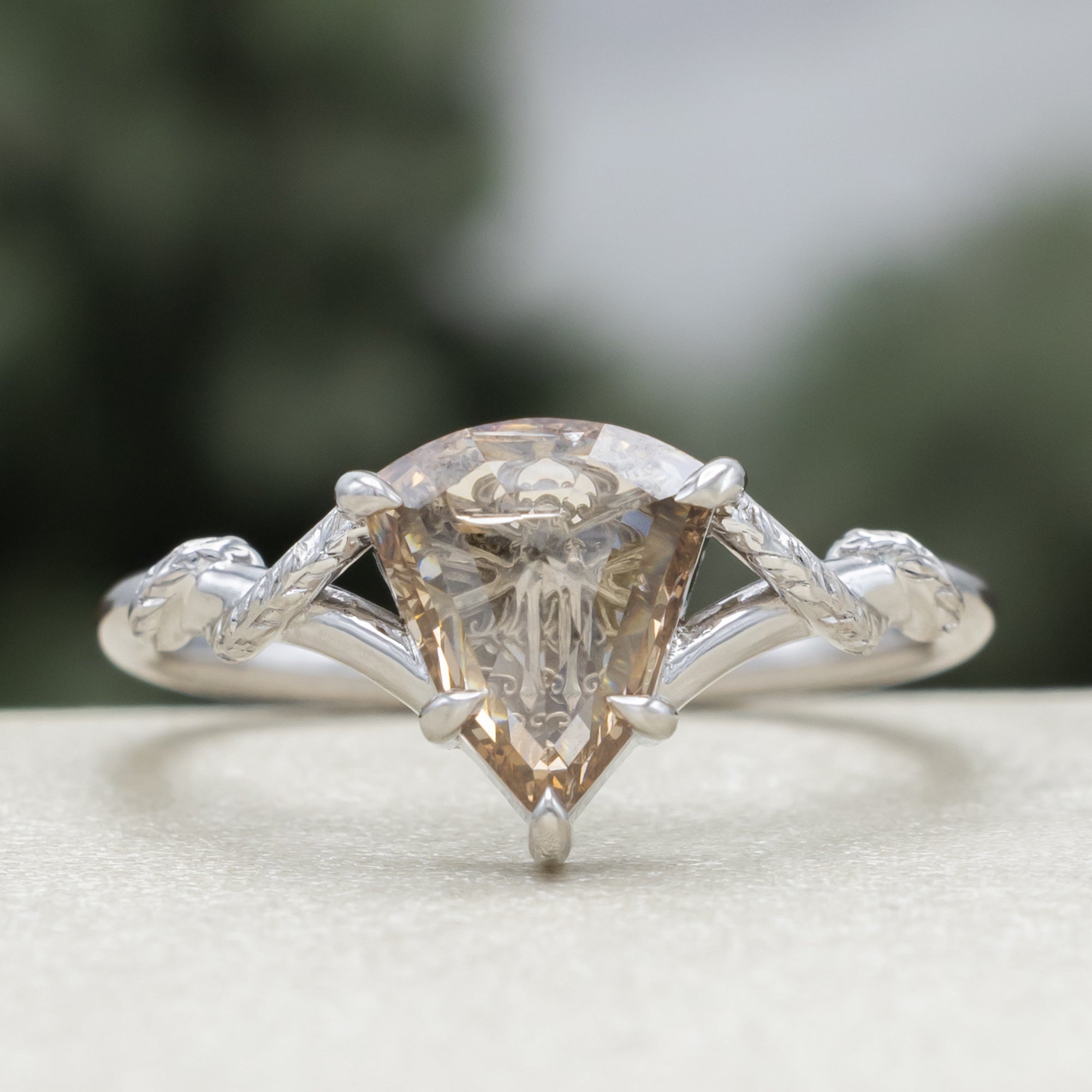 A white gold engagement ring featuring a shield-cut champagne diamond, with vine filigree twisting along the band against a soft blurred green background.