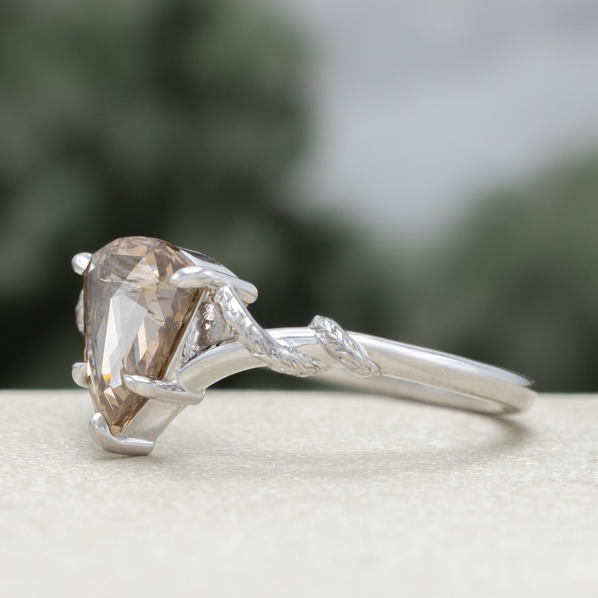 A white gold engagement ring featuring a shield-cut champagne diamond, with vine filigree twisting along the band against a soft blurred green background.