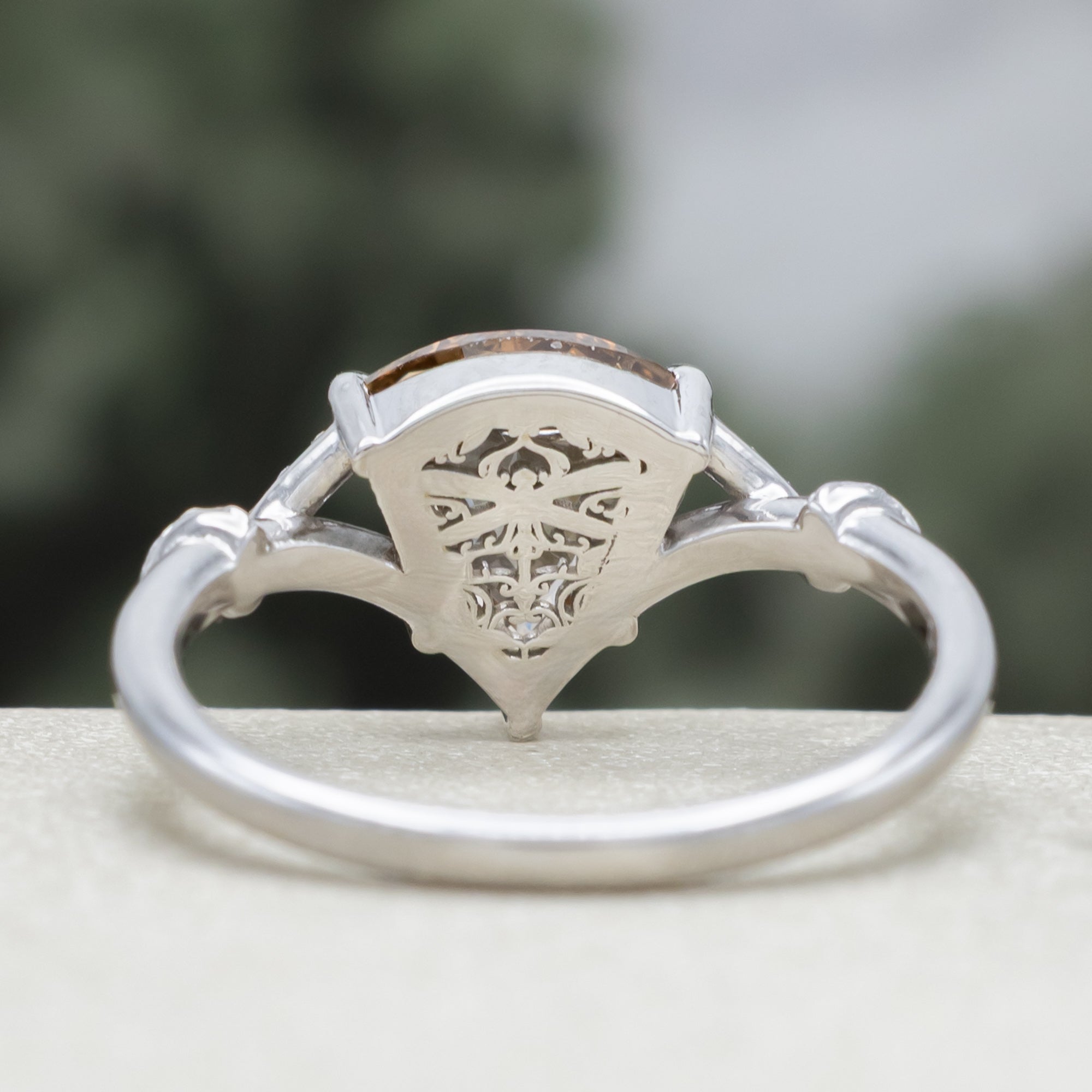 A white gold engagement ring featuring a shield-cut champagne diamond, with vine filigree twisting along the band against a soft blurred green background.